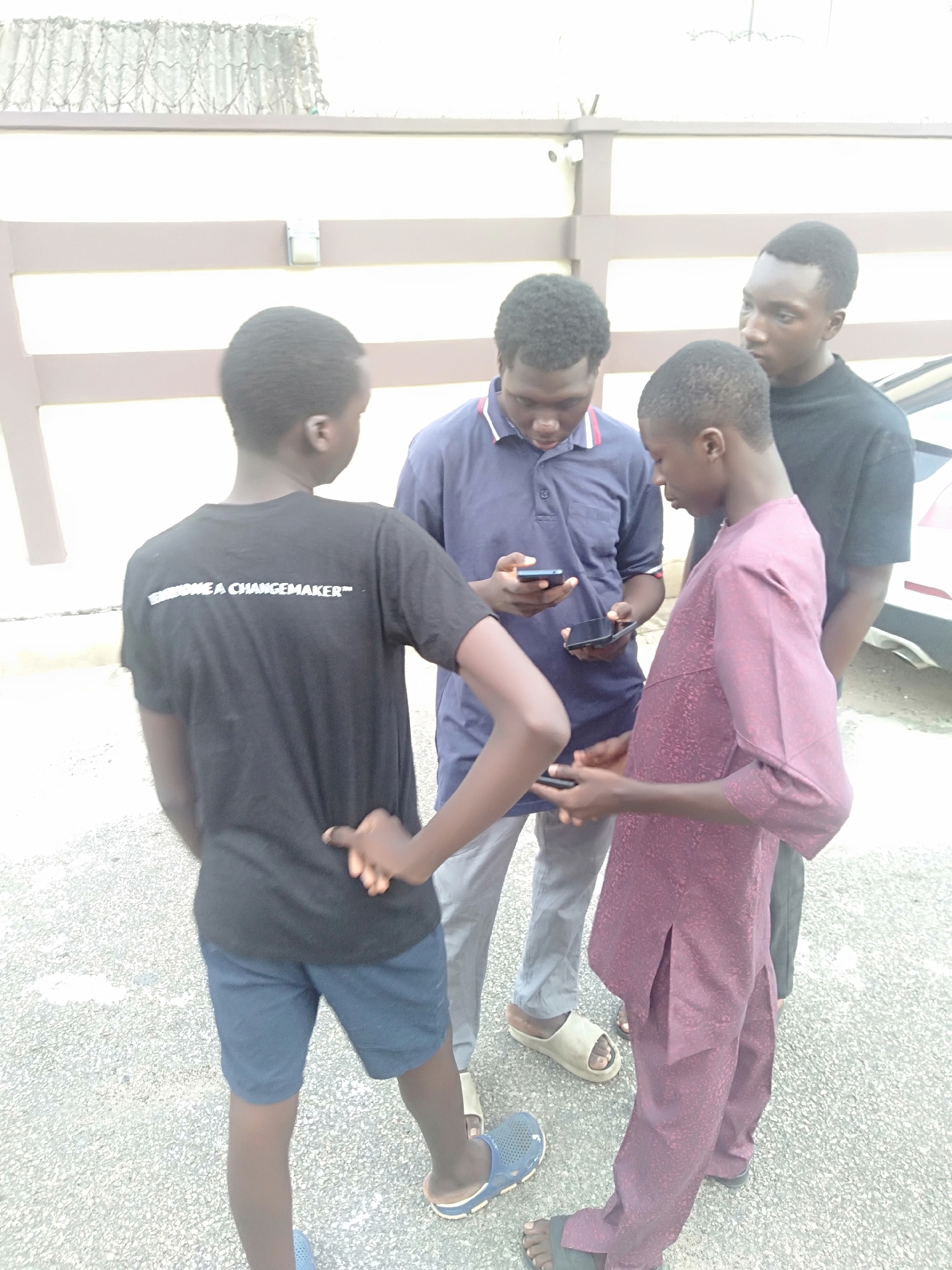 Four young males, appearing to be teenagers or young adults of African descent, are gathered in a casual huddle outdoors on a paved surface in Shangisha, Nigeria. Their attention is primarily directed downwards, with at least three individuals holding and looking at smartphones, suggesting they are sharing content, browsing, or engaged in a group activity on their devices.
The individual on the left, with his back to the viewer, wears a black t-shirt that prominently displays the text "BECOME A CHANGEMAKER™" across the shoulders, paired with blue shorts and blue slide sandals. Next to him, a male in a navy blue polo shirt, light grey trousers, and light beige slides is focused on his smartphone. A third boy wears a maroon patterned traditional-style tunic and trousers, also holding a smartphone and looking down, with black sandals on his feet. On the far right, partially visible, is a male in a black long-sleeved shirt, looking towards the group.
The scene features a light-colored, speckled paved ground and a building in the background with horizontal brown/beige slats. Barbed wire is visible along the top of a wall, and a white security camera is mounted on the building structure, indicating a secured property. Part of a light-colored car is visible on the far right. The bright, even lighting suggests it is daytime on a clear day.