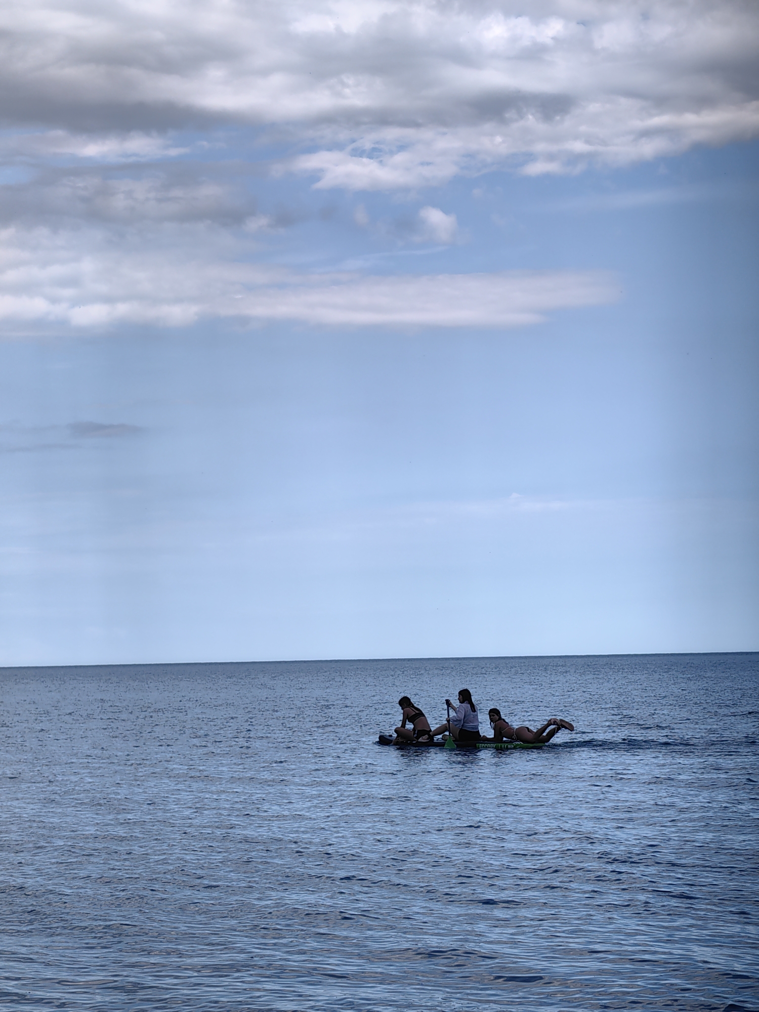 In the city of Utios, Ukraine, a serene body of water reflects a cloudy sky. Three individuals are present on a paddleboard. One person sits at the back, another is in the middle, and a third is lying down at the front. The water is calm with small ripples. The sky is a mix of blue and grey clouds, with the horizon clearly visible separating the water and the sky. The paddleboard is green and appears to be designed for multiple occupants.