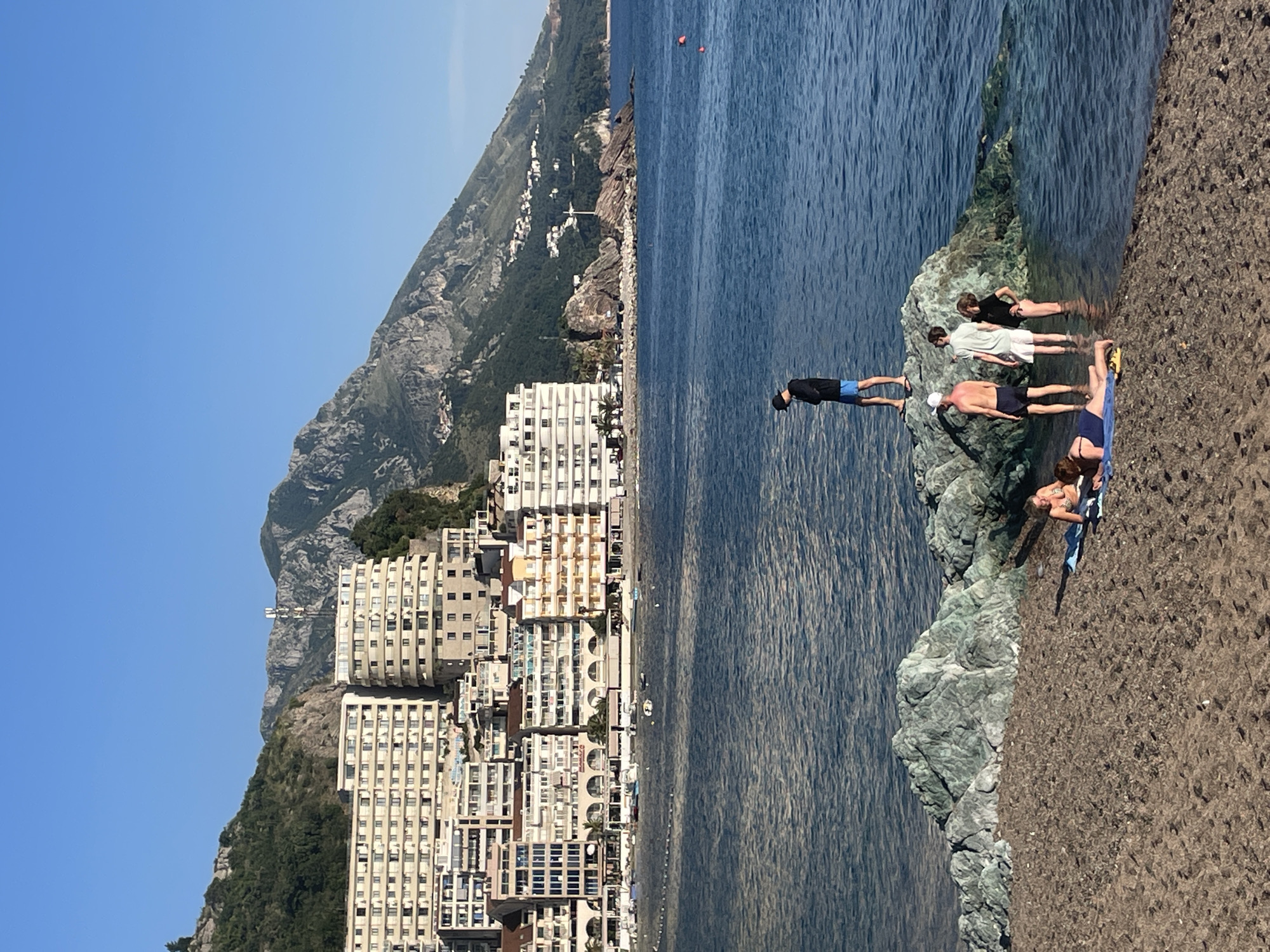 The image depicts a beach scene in Rafailovići, Montenegro, set against a backdrop of mountains and densely packed high-rise buildings. The buildings are a mixture of white and beige hues, densely clustered along the coastline. The mountains rise steeply behind the buildings, adding depth to the landscape.
In the foreground, people are present near the water's edge. Several individuals are standing on a rocky outcrop, with one person poised to dive into the sea. Other individuals are standing in the water, while a woman is lying on a towel on the beach. The water appears clear and blue. The beach itself is made of coarse sand. The time of day appears to be daytime, with clear blue skies.