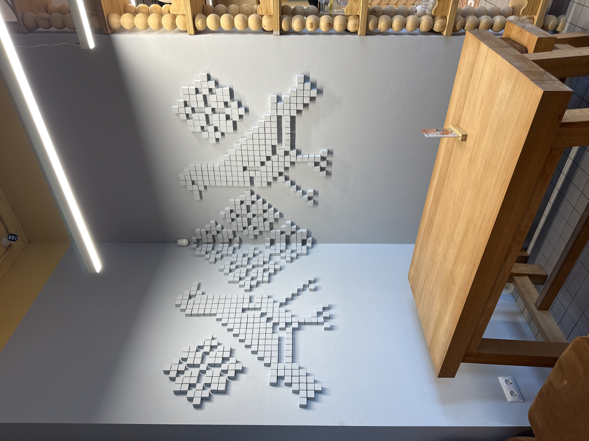 The image shows an interior scene with a wooden table in the foreground and a wall with decorative elements in the background. The table is made of light-colored wood and has a simple design. The wall features abstract pixelated designs that resemble birds or mythical creatures, created with white cubes. To the right of the wall art, there are wooden shelves with various items. A strip of light runs along the ceiling. The overall aesthetic suggests a modern and minimalist interior, possibly a cafe or restaurant. The location is identified as a cafe in Krasnodar, Russia.