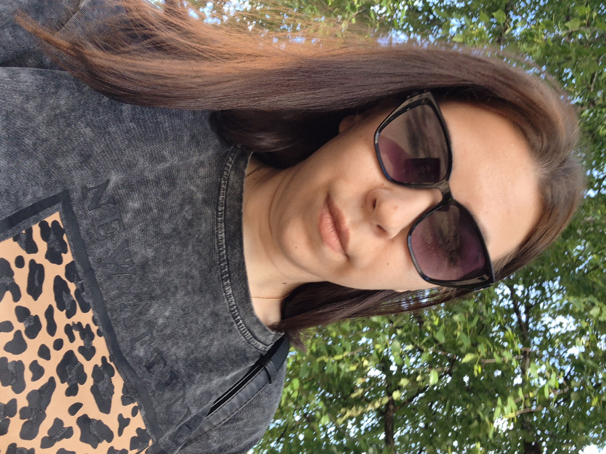 A tilted portrait shot features a person, likely female, outdoors in Chaikovsky, Russia, under bright daylight. The individual has long, dark brown hair, which flows across their shoulder and chest. They are wearing dark, square-framed sunglasses, partially obscuring their face, but showing light-colored skin and light pink lips. A delicate gold chain necklace is visible around their neck.
The person is wearing a charcoal grey sweatshirt with an acid-wash effect. On the left side of the sweatshirt, there is a large, light brown patch with a black leopard print pattern. Adjacent to this patch, on the grey fabric, vertically aligned letters are visible, including a clear "N" and "E", with hints of further text.
The background consists of blurred green foliage from trees, suggesting a natural outdoor setting such as a park or garden. The overall impression is a casual, sunlit portrait.