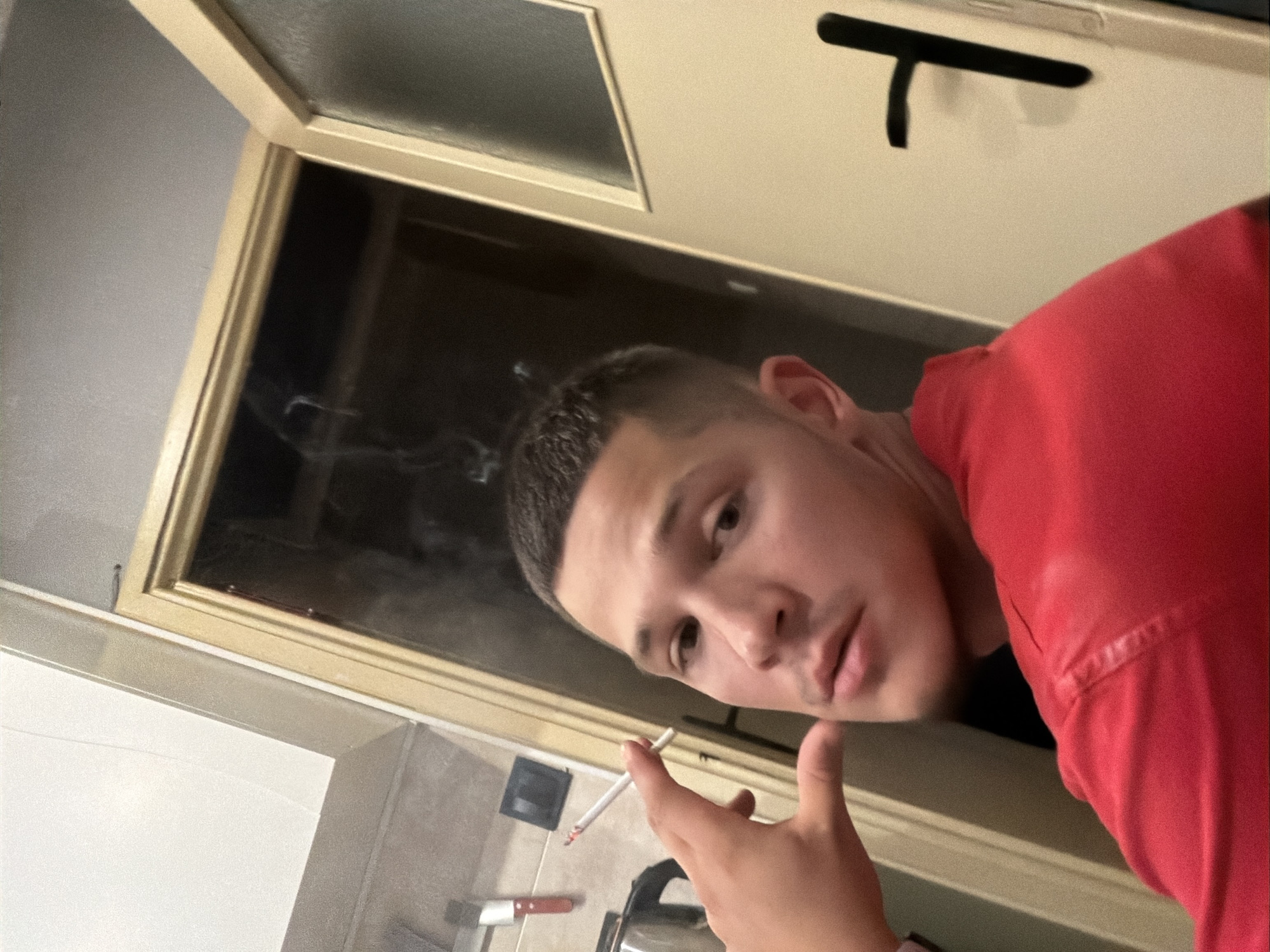 A young man with a short haircut is shown in a close-up, self-taken photograph. He is wearing a red shirt and holding a lit cigarette to his lips, with smoke rising from it. The background suggests an indoor environment, possibly a kitchen or entryway. A door with a black handle is visible to the right, and a kitchen counter with some utensils is present on the left. The lighting is dim, creating shadows. There are no specific indicators of the location within the city of Sofia, Bulgaria.