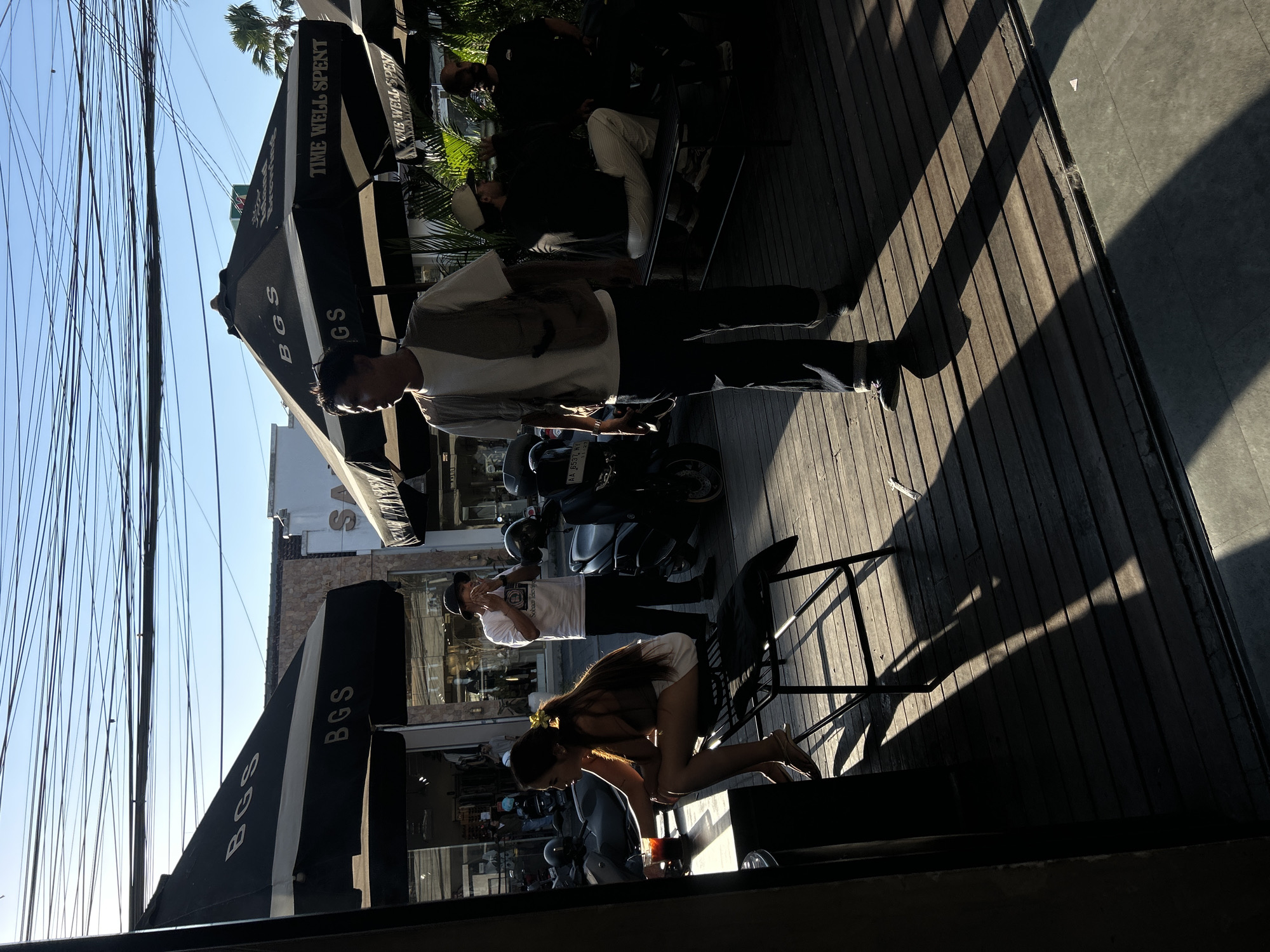 In North Kuta, Indonesia, a street scene captures a group of individuals in the late afternoon sun. The foreground shows a woman seated on a black metal chair at a wooden table, holding a drink. Next to her, a man in a white t-shirt and black pants stands smoking. To the right, another man in a vest and jeans stands on a wooden deck. In the background, under black umbrellas with "BGS" branding, other people are visible, some sitting on a bench. Scooters are parked on the left and right sides of the scene. The environment is characterized by a bright, sunny sky, overhead power lines, and wooden plank flooring.