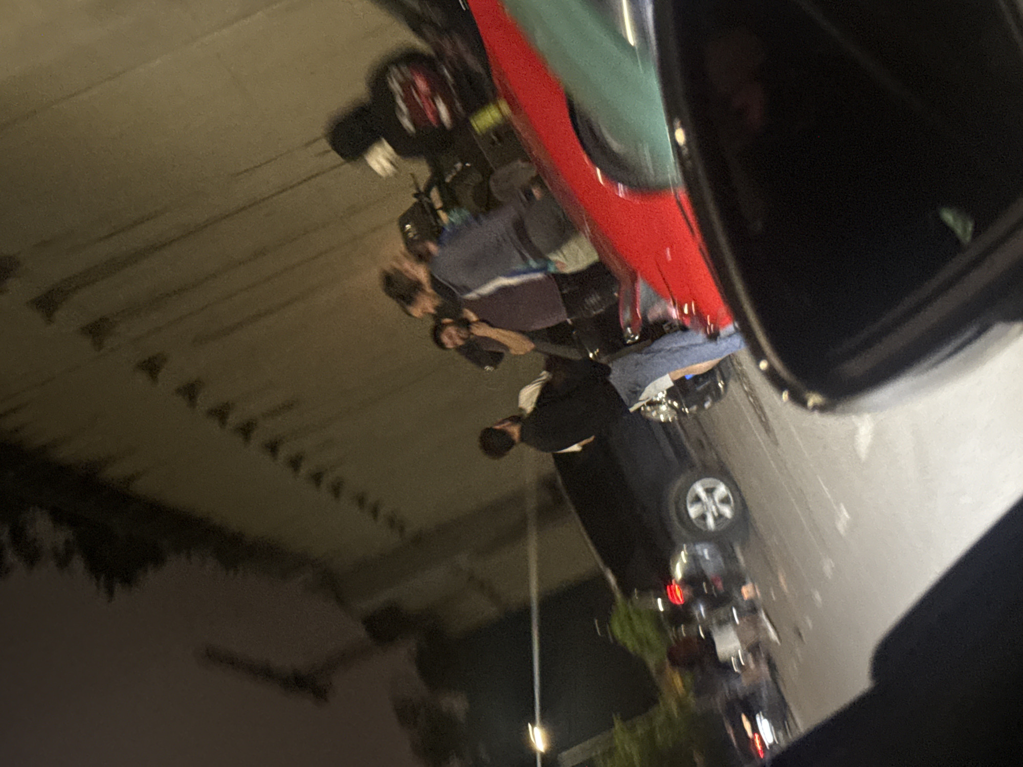 The image depicts a nighttime street scene in Varna, Bulgaria, captured from within a vehicle at a slight upward and rightward angle. Multiple individuals, primarily adult males, are gathered on the asphalt street and sidewalk area.
In the mid-ground, a dark-colored SUV, possibly black, is parked with its front bumper facing right. To its left, a male wearing a dark long-sleeved shirt and blue jeans stands facing right, appearing to interact with a group of approximately three other males to his immediate right. This group shows varied expressions, with some appearing to smile or converse. Further to the right, another individual in a dark hoodie with a lighter-colored interior or undershirt is partially visible next to a red sports car.
The red sports car occupies a significant portion of the right mid-ground and foreground, presenting a partial view of its driver's side. A teal or dark green vehicle is partially visible behind the red car. In the bottom left, a dark car interior element, possibly a door panel, is visible, indicating the perspective of the shot. Another vehicle's red taillights are faintly visible further down the street on the left.
The background is dominated by a tall, light-colored concrete or stone wall, possibly part of a building or overpass structure. This wall features multiple rows of small, dark, triangular or pointed protrusions or shadow patterns along its surface. A bright streetlight is visible on the left side of the frame, illuminating portions of the street and background. The overall lighting is artificial and dim, characteristic of nighttime. The image exhibits motion blur, particularly affecting some of the people and the general sharpness of the scene, suggesting movement during capture or subjects in motion.