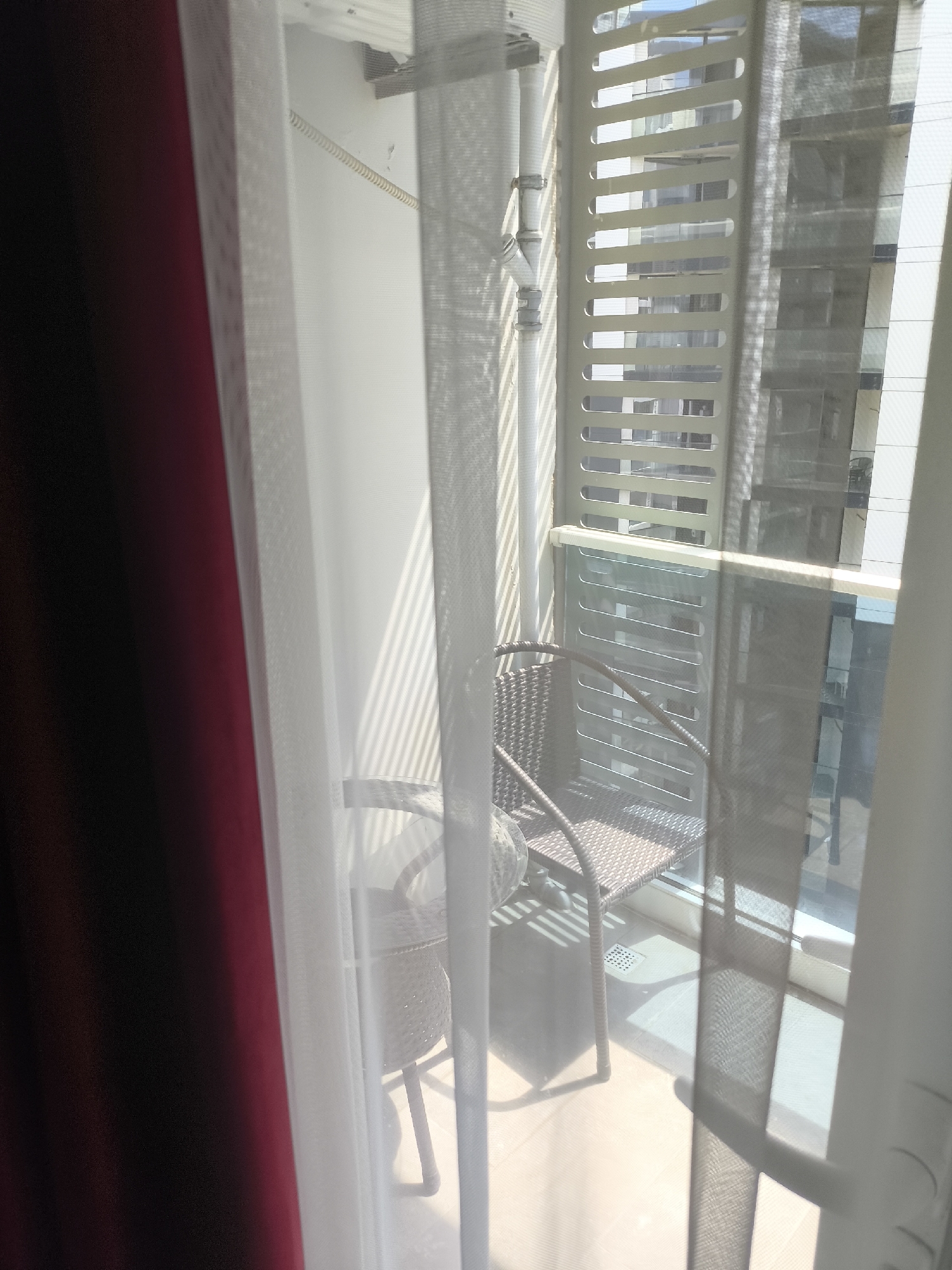 The image depicts a balcony area, likely in Batumi, Georgia, viewed through sheer curtains. A single chair made of woven material, with a cushioned seat, is visible on the tiled balcony floor. The chair is positioned in front of a slatted metal partition that separates it from the outside. Sunlight casts striped shadows across the balcony. Beyond the partition, a modern building with multiple windows is reflected. A dark red curtain is present on the left side of the frame, and sheer white curtains partially obscure the view. A drain grate is visible in the floor tiling. The scene appears to be an unoccupied outdoor seating area.