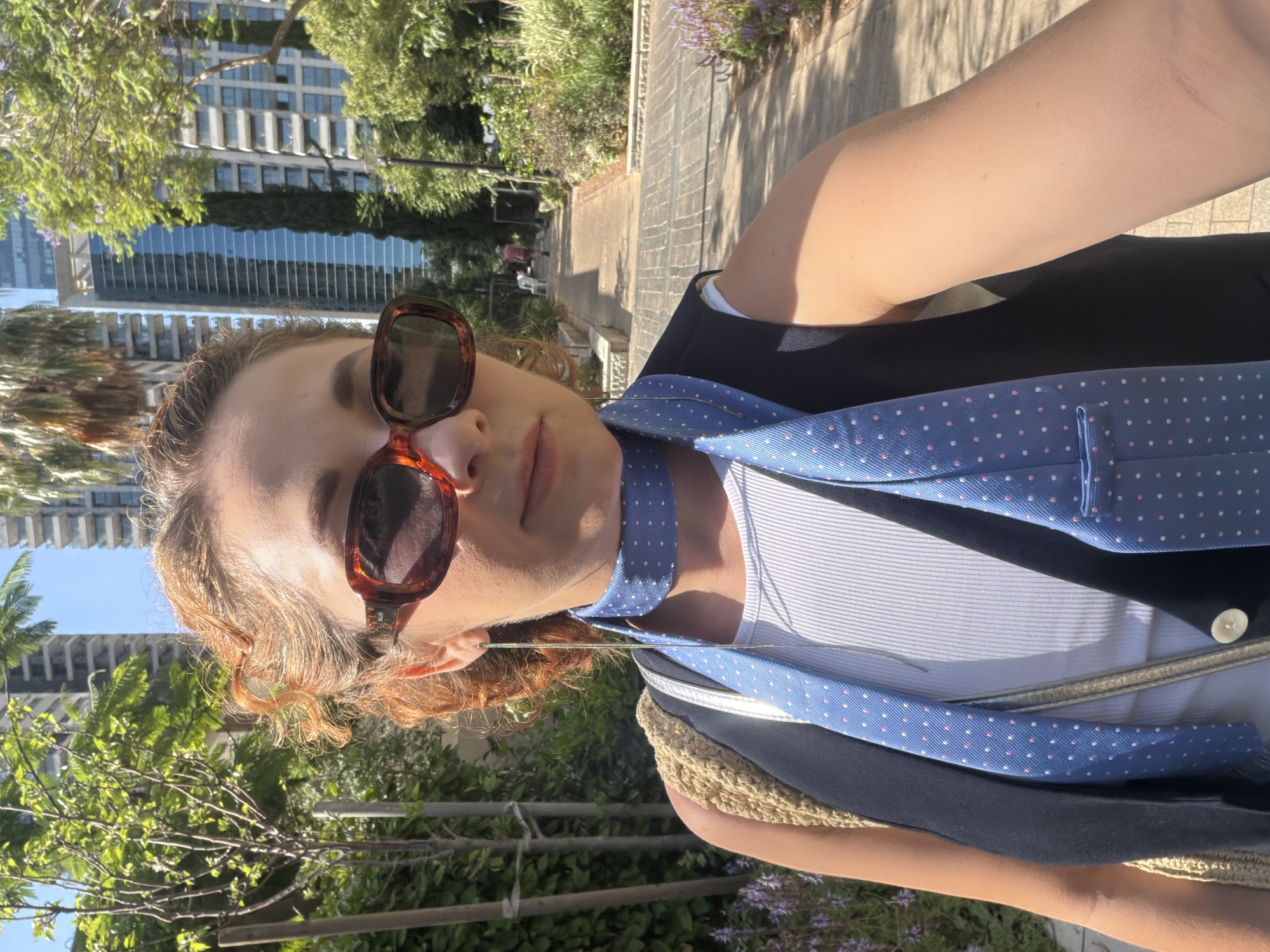 A medium shot captures a woman from the chest up, wearing sunglasses and a blue tie with pink polka dots. She has auburn, curly hair pulled back and is looking directly at the camera. The background reveals a sunlit urban park setting in Tel Aviv, Israel, with lush green trees, modern high-rise buildings, and a paved walkway. The woman is holding a light brown woven bag over her left shoulder.