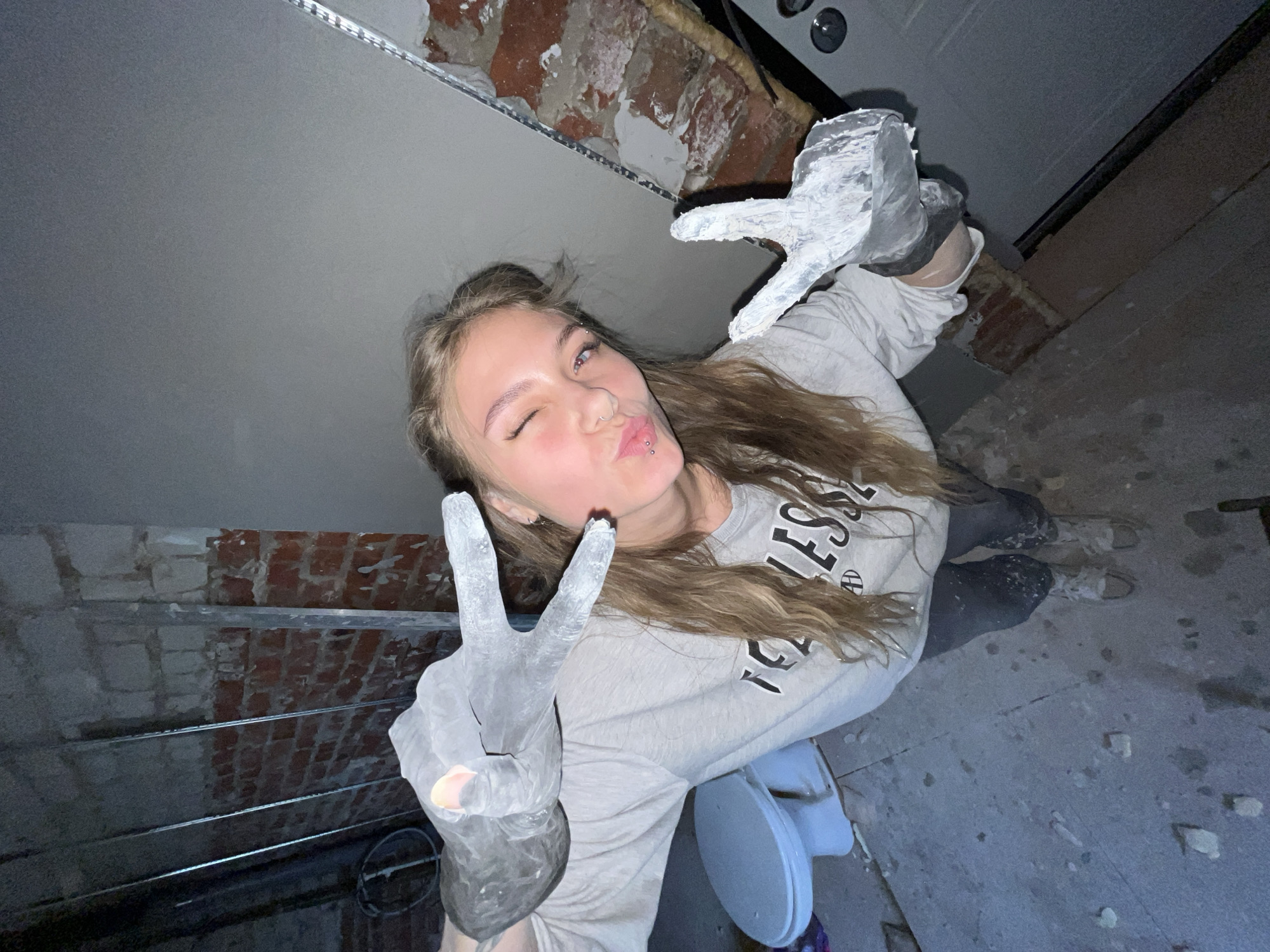 A young woman with blonde hair is making a peace sign with both of her hands, which are covered in gray-white material. She is winking and has her lips puckered. The woman is wearing a light-colored sweatshirt with the word "BLESS" printed on it. The background appears to be an indoor construction site, with exposed brick walls and a newly installed ceiling. The lighting is dim, possibly from artificial sources. There are visible signs of construction work, such as building materials. The city is not discernible from the image.