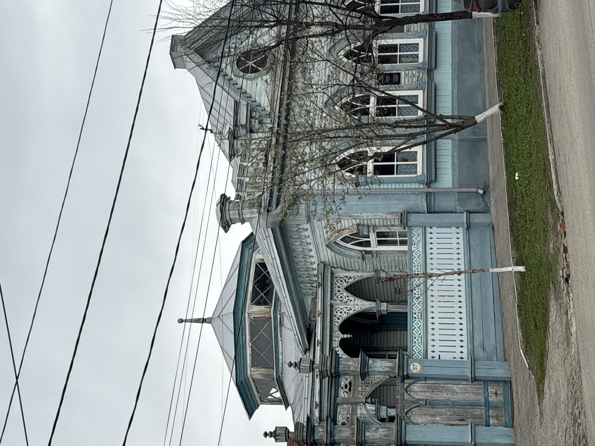 A color photograph shows an ornate, two-story, light-blue wooden building at 57 Radishcheva Street, Ulyanovsk, Russia. The building features elaborate woodwork, arched windows, and decorative trim. A bare tree stands in front of the building. Overhead power lines are visible against a cloudy sky. A narrow strip of grass and a paved road are to the right of the building.