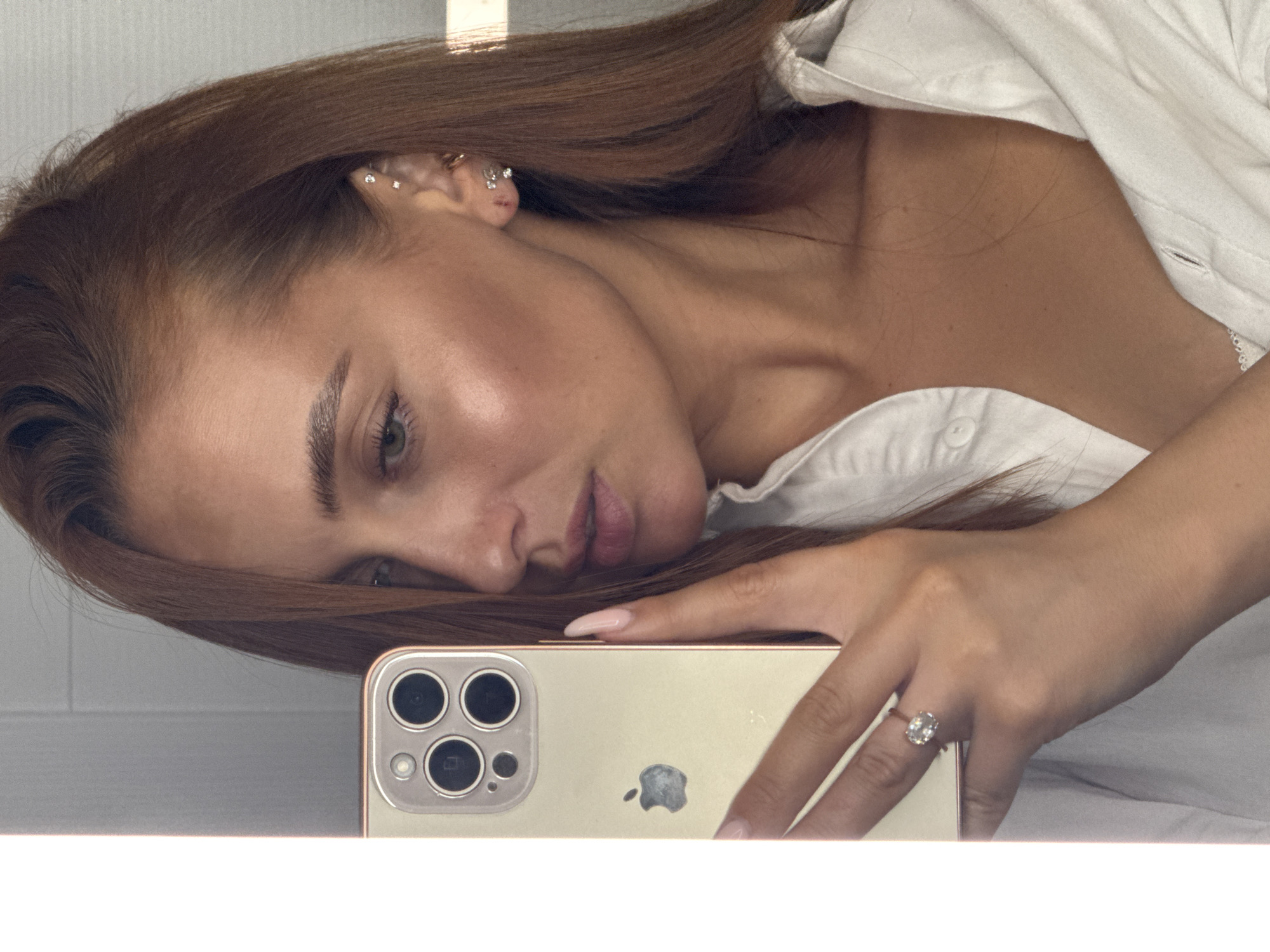 The image shows a close-up, top-down, sideways shot of a woman lying down and holding a smartphone. The woman has long, brown hair that is partially obscuring her face. She is wearing a white, collared shirt that is unbuttoned, revealing her neck and chest. Her face is turned towards the camera, and her eyes are partially closed. She has delicate jewelry on her ear. She is wearing a ring with a large oval stone on her left ring finger. The smartphone is a light-colored iPhone, positioned horizontally in her hand, with the camera lenses facing the viewer. The woman's fingernails are painted a light pink. The background appears to be a light-colored, possibly tiled surface. There is a bright light source casting a shadow on the woman's hair. The lighting suggests it is daytime, and the indoor setting is likely a bedroom or similar private space. The woman's expression is relaxed and possibly contemplative.