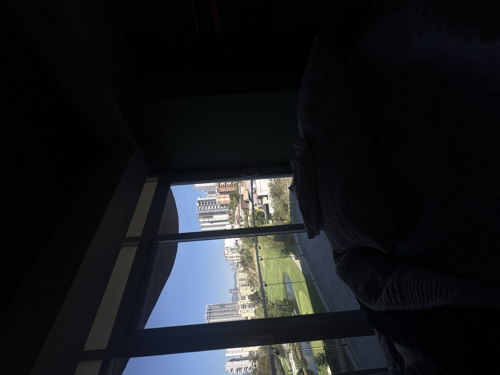 The image shows a view through a window in a darkened room, looking out at a city landscape in Huixquilucan de Degollado, Mexico. The window is framed by dark walls and ceiling, with a portion of a large, dark object (possibly a piece of furniture or drapery) obscuring the right side of the view. The cityscape visible through the window includes a golf course and buildings of varying heights, suggesting a densely populated urban area. The sky is clear and blue, indicating sunny weather conditions.