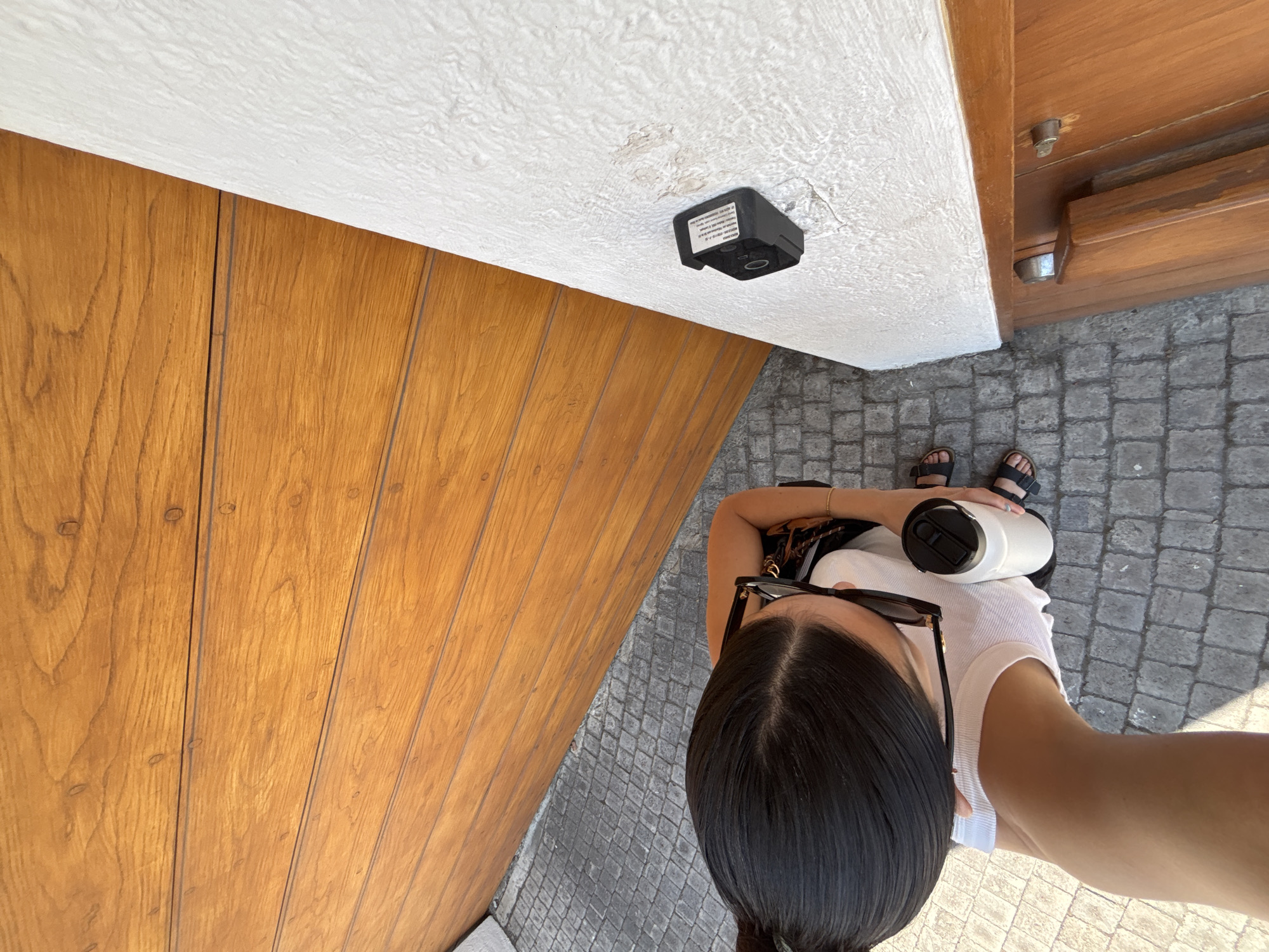 An overhead photograph captures a person standing at an entrance or passageway, characterized by a mix of natural materials and a bright, outdoor setting.
The main subject is a person, likely female, seen from the top of their head down to their feet. They have dark hair parted centrally, with black sunglasses resting on top of their head. They are wearing a white short-sleeved top and black sandals. In their right hand, they hold a white insulated water bottle with a black lid. A portion of a patterned bag strap is visible on their left shoulder.
The immediate surroundings feature a prominent wooden wall or door on the left, composed of vertical panels with visible small, circular fasteners. Above, a white, heavily textured ceiling or overhang extends, from which a small, black, rectangular electronic device is mounted. This device has an unreadable label with tiny text on its side. The ground beneath the person's feet is a pattern of grey cobblestones, suggesting an outdoor or semi-outdoor public space. To the far right, another section of wooden structure with metallic hardware is visible.
The scene is brightly lit, indicating it is daytime. The combination of wooden architectural elements and cobblestone paving evokes a traditional or historic ambiance, consistent with locations in San Bernardino Tlaxcalancingo, Mexico. The high vantage point suggests the photo was taken as a downward-facing selfie or by holding the camera directly overhead. No specific activity beyond standing and holding the bottle is clearly depicted, nor are any emotions discernible.