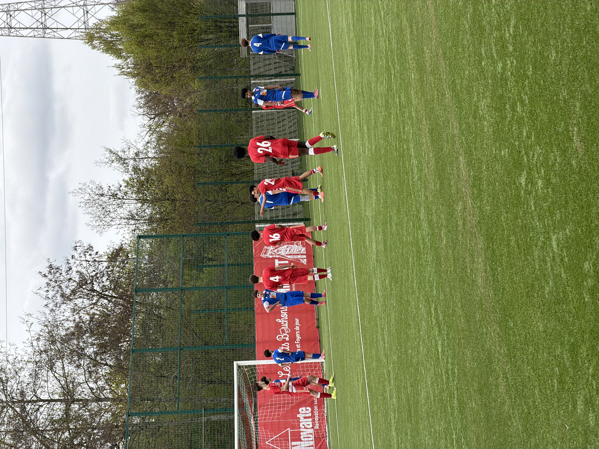 The image shows a soccer field in Esch-sur-Alzette, Luxembourg. The field is green with evenly spaced white lines. A line of soccer players is visible near one of the goals. The players are wearing red and blue uniforms. The team in red is facing a free kick. A red banner with white letters partially covers the goal. A chain link fence runs along the side of the field, with trees behind it. Power lines are visible in the background. The sky is overcast.