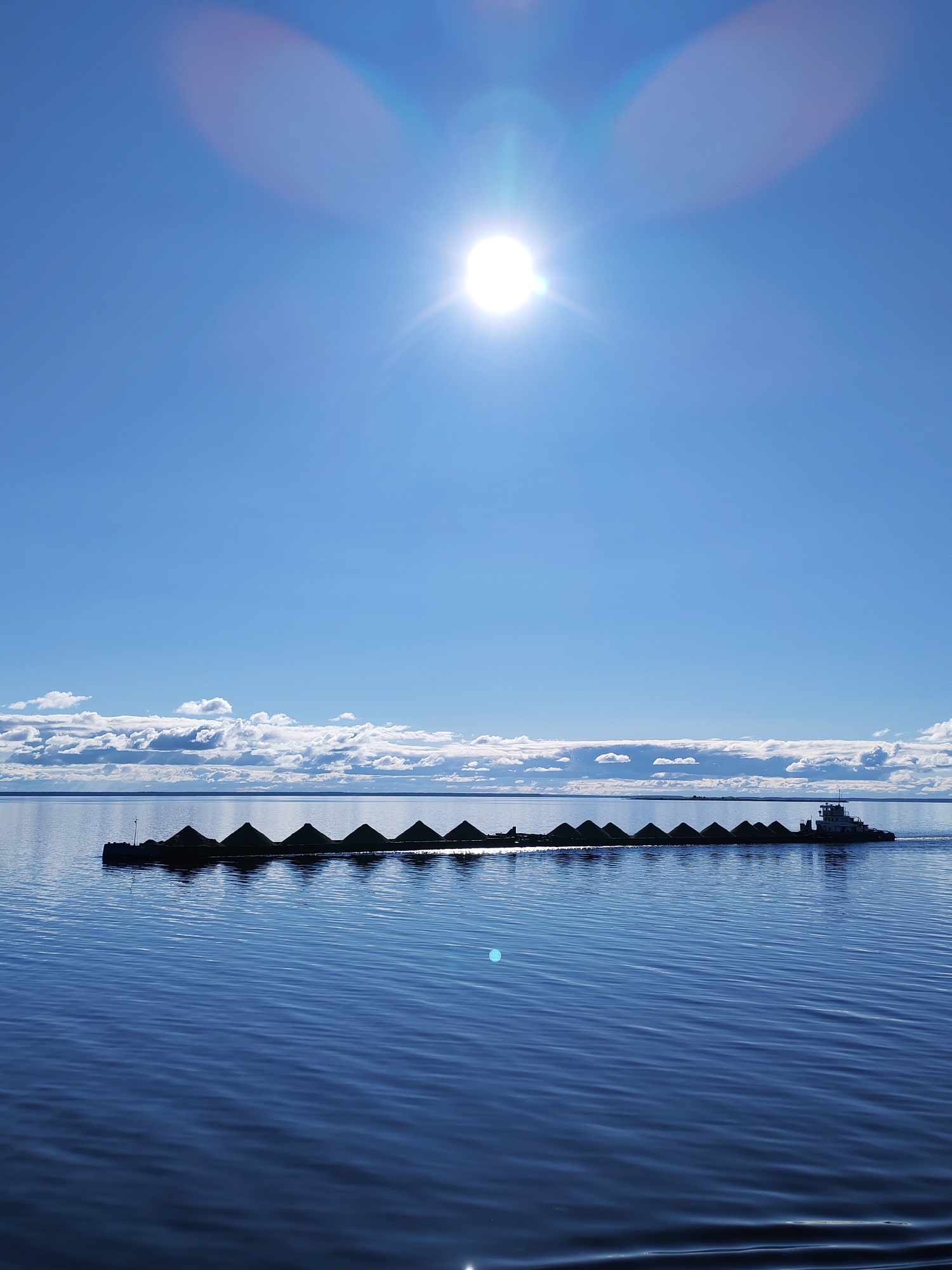 A photograph captures a long barge, laden with conical mounds of dark material, being pushed across a large body of water by a tugboat. The scene is brightly lit, with the sun positioned high in the blue sky. The water is a deep blue, reflecting the sky and the sun's glare, and ripples across its surface. Along the horizon, white clouds create a distinct separation between the sky and the water. The tugboat is a light color and is located at the rear of the barge. This image was taken in Mikhalevo, Russia.