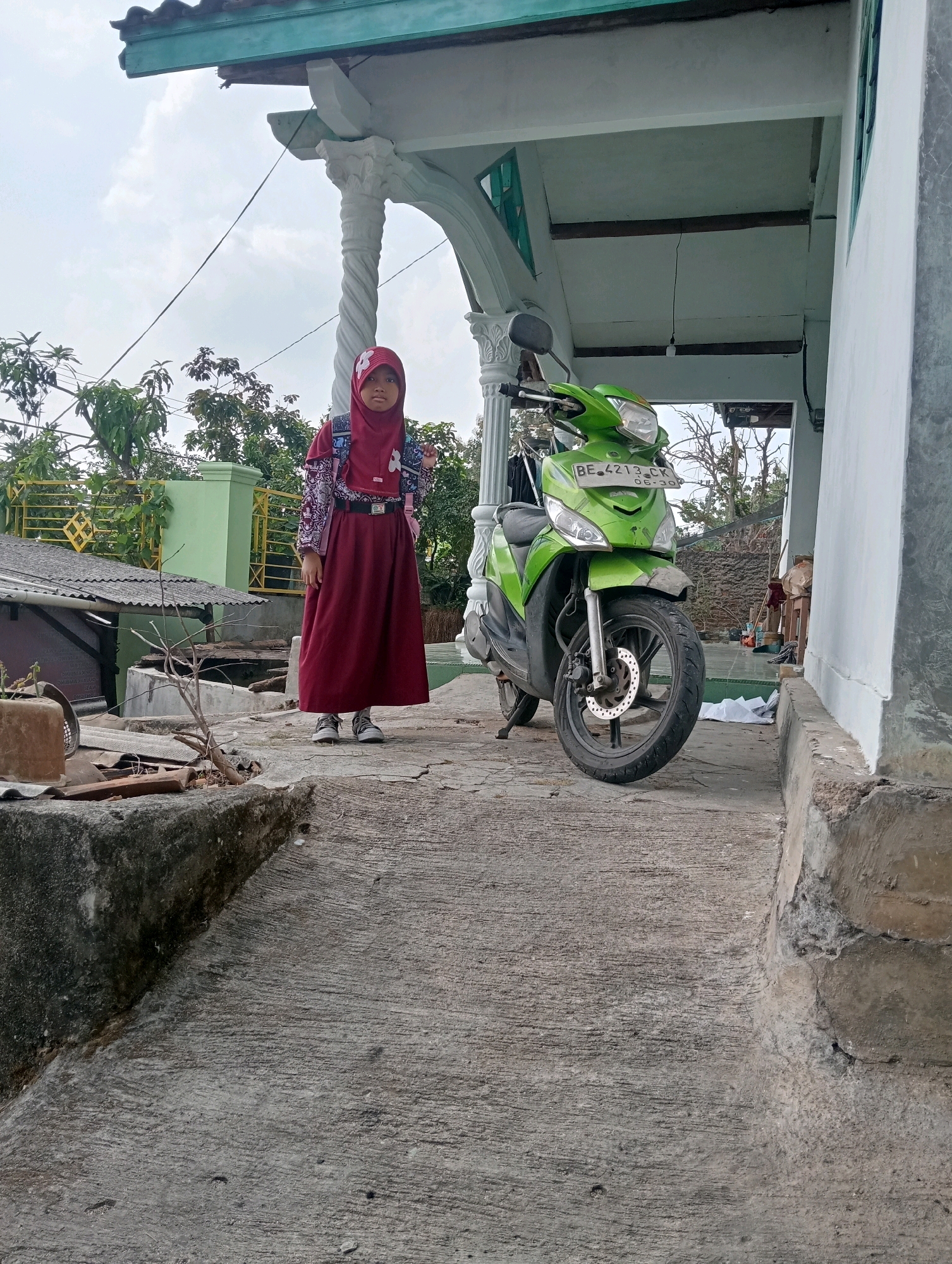A young girl wearing a maroon hijab, school uniform, and backpack stands on a concrete path next to a green scooter. The girl is looking directly at the camera. The scooter is parked under a covered outdoor area with a white pillar and green trim. In the background, there are trees and a building. The scene appears to be in a residential area in Bandar Lampung, Indonesia.