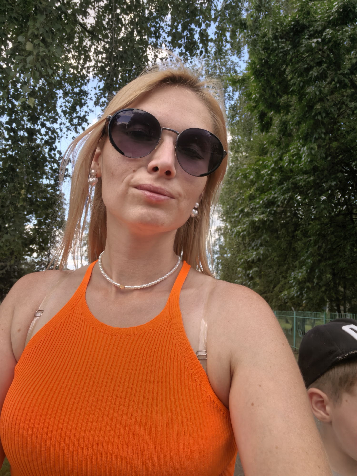 The image is a selfie taken outdoors in a city park with lush green trees and a bright blue sky. The primary subject is a fair-skinned woman with blonde hair, wearing large, dark-rimmed sunglasses and an orange ribbed tank top. She is also wearing a pearl necklace with a small gold heart pendant and heart-shaped pearl earrings. The woman has a slight smile and her head is tilted. To her right, a young boy wearing a black baseball cap with a white logo is partially visible. The background suggests a public outdoor space, possibly a park in Sterlitamak, Russia, given the context provided, with hints of fencing and other greenery. The lighting indicates it is daytime, likely sunny.