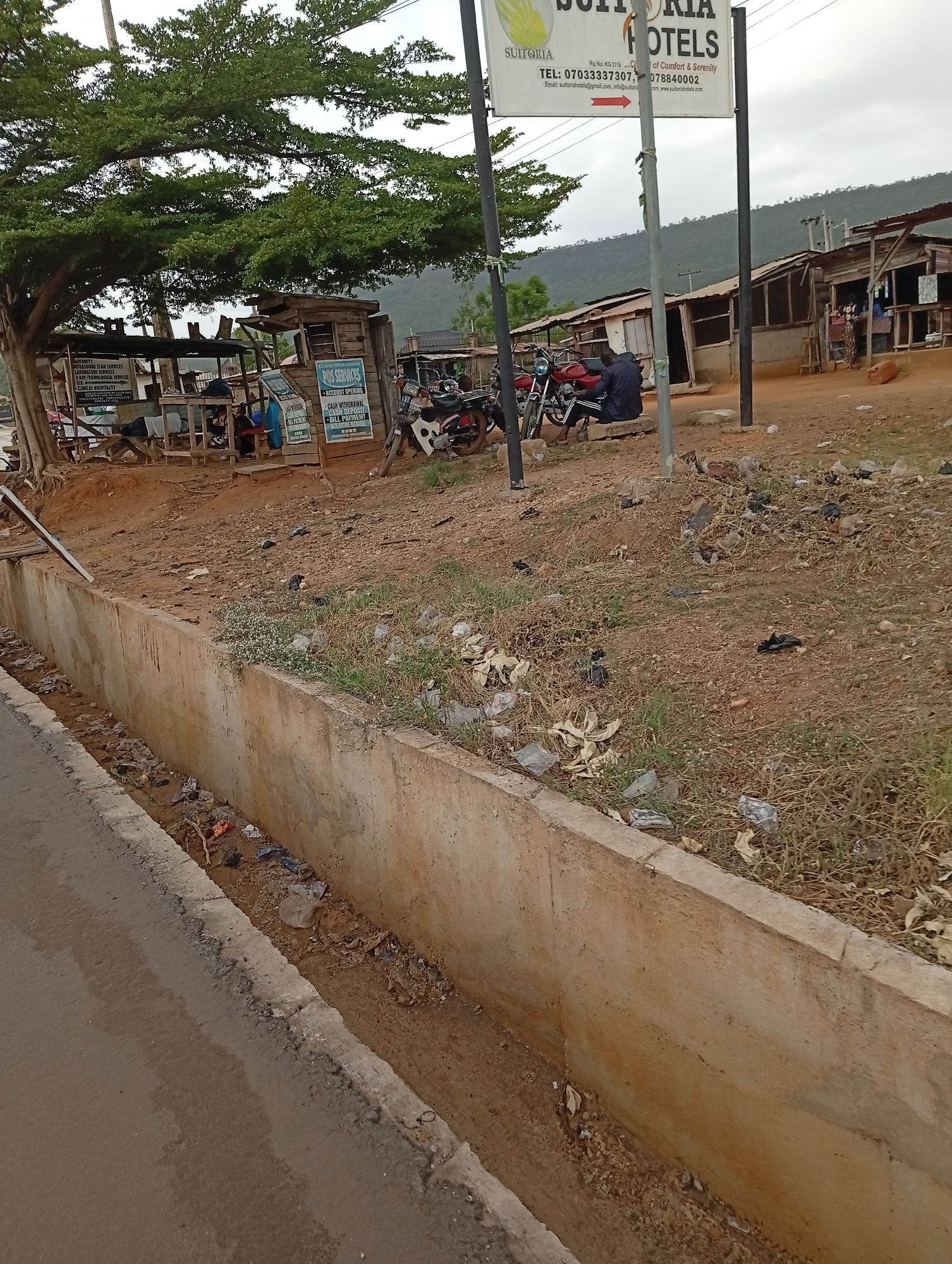The image depicts a street scene in Lokoja, Nigeria. A concrete drainage ditch runs along the road in the foreground, containing trash. Beyond the ditch is a dirt area with sparse vegetation and scattered trash. Further back, there are several small, makeshift shops or stalls, one partially shaded by a large tree. There are motorcycles parked near the shops. A sign for "Suitoria Hotels" is visible, advertising comfort and serenity. In the background, there is a view of hills or mountains. One person is sitting on a bench. The OCR reads: "MATERNITY ULTRASOUND SCAN SERVICES, LABORATORY SERVICES, X-RAY/RADIOLOGICAL SERVICES, ECG, CLINICAL HOSPITALITY", "POS SERVICES - ACCOUNT OPENING - CASH WITHDRAWAL - CASH DEPOSIT - BILL PAYMENT", "SUITORIA HOTELS of Comfort & Serenity Rg No. KG 3119C TEL: 07033337307, 078840002 Email: suitoriahotels@gmail.com, info@suitona com, www.suitoriahotels.com"