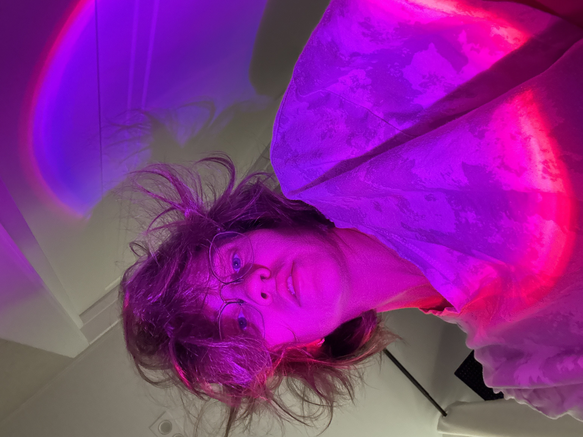 An indoor portrait features a young person, possibly female, looking directly at the camera. The image is rotated counter-clockwise, suggesting a low-angle shot or the subject lying down. The person has messy brown hair, illuminated vibrantly with purple and pink light, and wears clear-rimmed glasses, revealing blue eyes. Their expression is soft and neutral, with a slight hint of a smile.
The scene is dominated by striking artificial lighting in shades of purple and hot pink, creating a dynamic and atmospheric ambiance. These lights cast strong, colorful hues across the subject's face, hair, and a light-colored garment they are wearing, which appears to be a shirt or a draped fabric. A distinct arc of bright purple and red light is visible on the wall to the upper left, suggesting the use of a special lamp (e.g., a sunset lamp).
The background consists of a plain wall, partially obscured by the colorful light and shadows. Hints of a ceiling and possibly a dark rectangular electronic device are visible in the lower right portion of the rotated image. The heavy reliance on artificial lighting suggests the photo was taken during the evening or night. No text is visible in the image. The location context is Pervomaysky, Russia.