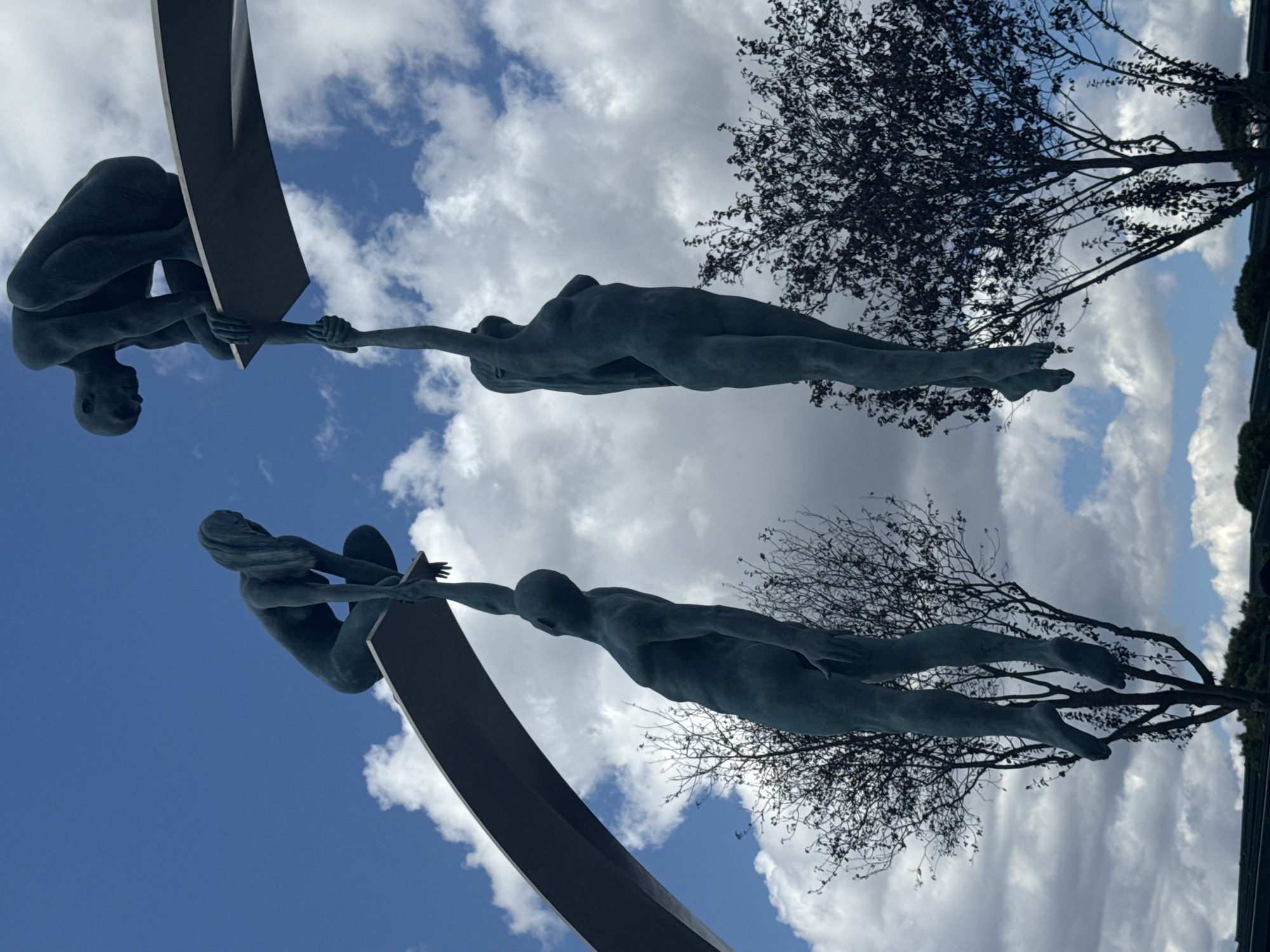 The image depicts a bronze sculpture located in Krasnodar, Russia. The sculpture, titled "The Ascent" by artist Nikolai Kondratenko, features four human figures in a dynamic pose against a backdrop of a blue sky with white clouds. Two figures are perched on a diving board-like structure, one in a crouched position reaching down, and the other in a kneeling position reaching up. Below, two other figures are suspended, one reaching up to grasp the hand of the figure above, and the other hanging limply. The sculpture is surrounded by trees, some bare and others with dark foliage. The overall scene conveys a sense of struggle, support, and aspiration.