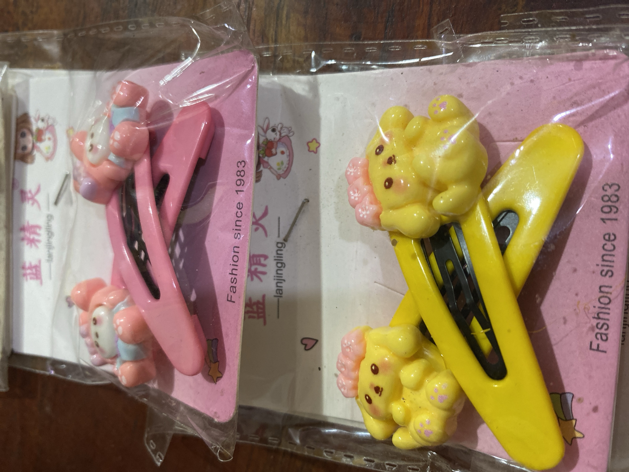 This image displays two sets of character-themed hair clips. The top set features pink hair clips adorned with a white and pink character resembling a bear or rabbit, wearing a light blue outfit. The second set, positioned below the first, showcases yellow hair clips with a yellow character that appears to be a dog with large ears and pink accents. Both sets are packaged on pink cardstock with text. The text on the top card reads "Fashion since 1983" and "lanjingling". The bottom card also has "Fashion since 1983" visible. The items are laid out on a wooden surface. The image's perspective suggests it was taken from above, looking down at the products. The lighting is natural, casting some shadows. There is no visible information within the image that indicates a specific location in Ta', Indonesia.