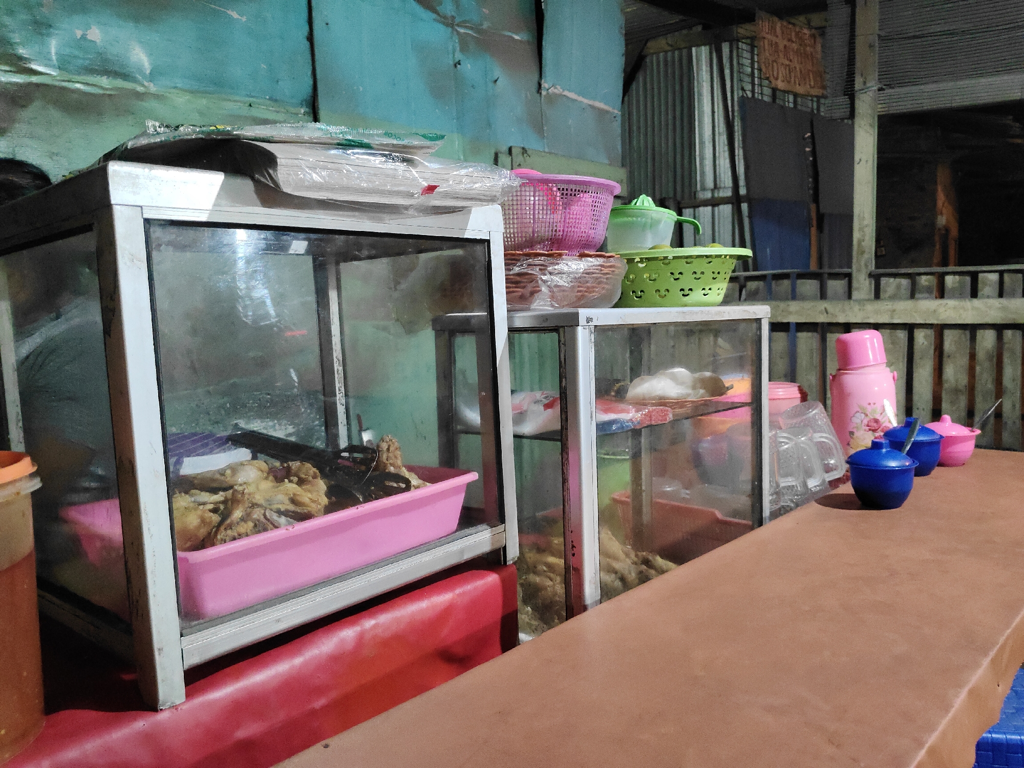 This is a medium shot, eye-level, outdoor night photograph of a food stall in Watukosek, Indonesia. The foreground is dominated by a brown table, with a red padded bench visible beneath a glass display case on the left. The display case contains fried food in a pink plastic tray. Behind it, on a shelf, are stacks of paper products wrapped in plastic.
To the right of the first display case is another glass display case, also with food visible inside. On top of this second case are several plastic baskets, a manual juicer, and some round, brown items in plastic. Further to the right, on the brown table, are a pink thermos, a blue bowl with a spoon, and a pink bowl with a spoon. The background features a wooden fence and corrugated metal walls. The overall lighting is dim, suggesting nighttime. There are no people clearly visible in the image.