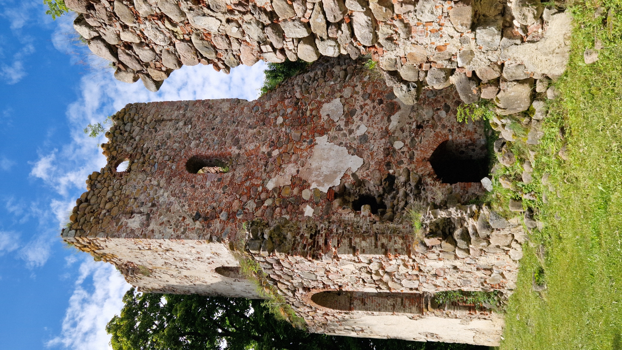 The image shows the ruins of the castle of Embūte in Embūte, Latvia. The ruins are composed of stone and brick, with some sections of the walls still standing tall. There are several arched openings and windows visible in the structure. Vegetation, including grass and trees, surrounds the ruins, and a blue sky with clouds is visible in the background. The scene is a historical ruin with no people present.