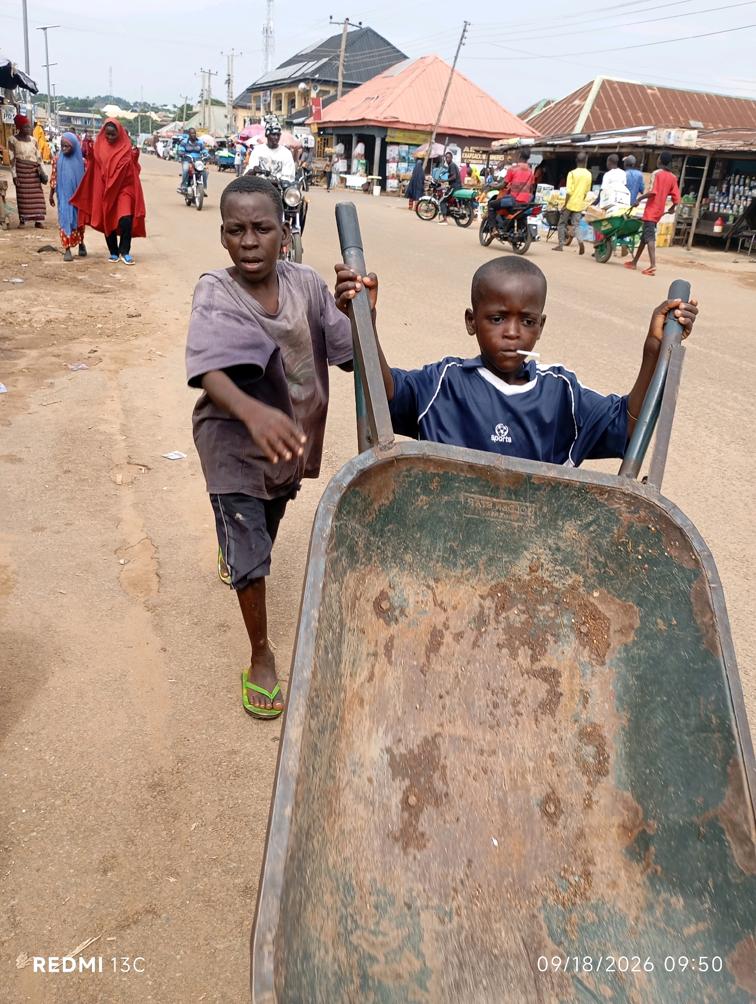 Two young boys are pushing a rusty wheelbarrow on a dusty road in Shendam, Nigeria. The boy in the foreground is wearing a blue and white athletic shirt and has a lollipop in his mouth. The boy behind him is wearing a faded purple t-shirt and dark shorts. They are walking towards the viewer, with the wheelbarrow taking up most of the right side of the frame. In the background, there are market stalls, people walking, and motorcycles. The sky is overcast. The road appears to be unpaved, with loose gravel and dirt. There are some pieces of trash scattered on the ground. The buildings in the background are simple structures, some with corrugated metal roofs. The scene suggests a bustling marketplace on a typical day.