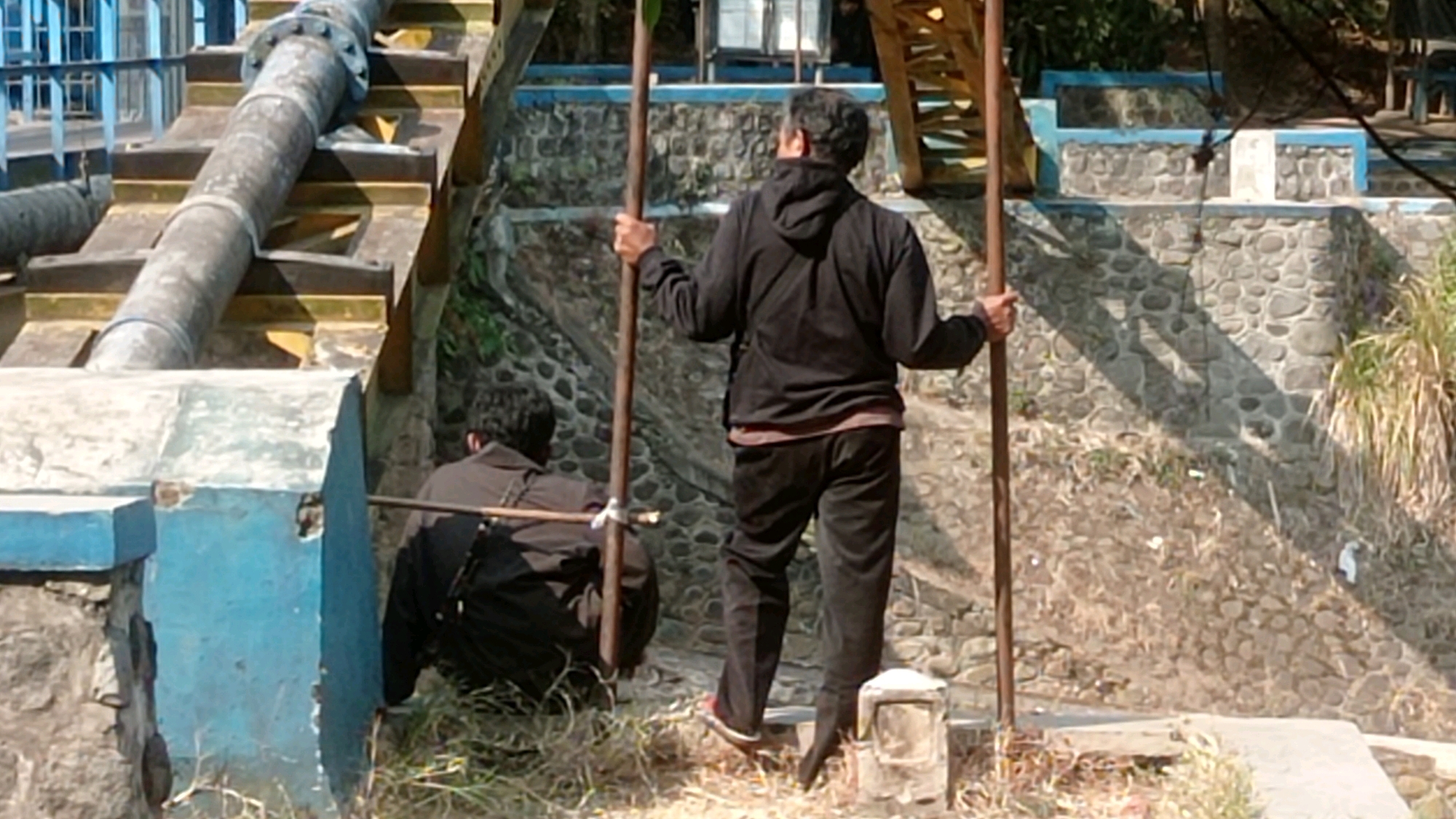 Two individuals are positioned outdoors near a water management structure. The person in the foreground, facing away from the camera, wears a black hooded jacket and dark pants, holding two wooden poles. To the left and slightly behind, another person, also facing away, is crouched down wearing a dark jacket and pants, holding a wooden pole that extends across their body. The background features a concrete and stone embankment with a large pipe running diagonally upwards. A blue structure is visible on the left, and foliage is present on the right. The scene appears to be in a rural or semi-urban area with evidence of water infrastructure.