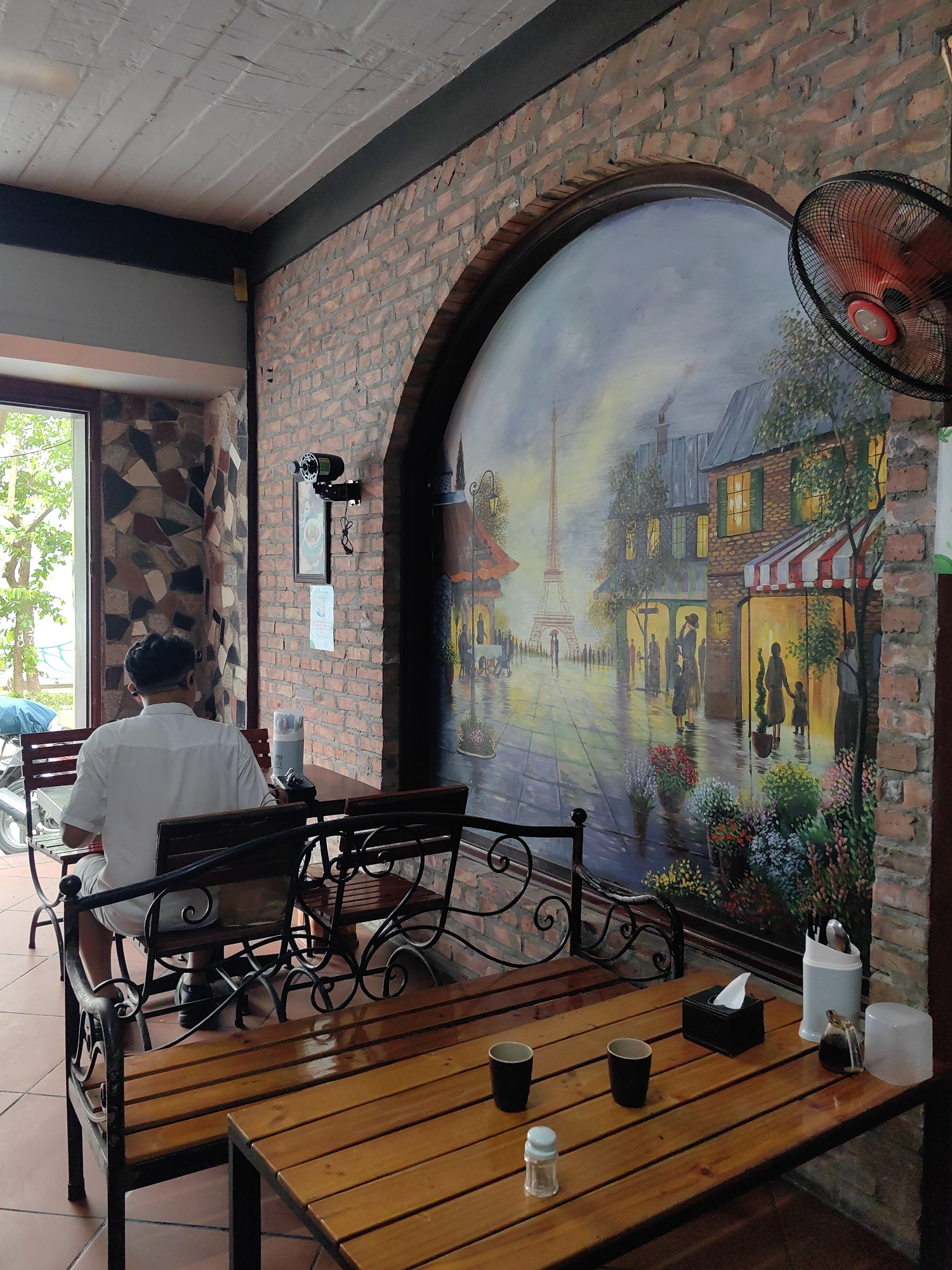The image depicts the interior of a café or restaurant in Hà Nội, Vietnam, characterized by exposed brick walls and decorative elements. A man in a white shirt sits at a table with wrought-iron chairs and is facing away from the camera. On the brick wall is a large mural depicting a Parisian street scene, complete with the Eiffel Tower in the background. Above the man, a surveillance camera is mounted on the wall. A small sign is seen on the brick wall. A red fan is visible in the top right corner.
On the foreground table, made of wood, there are cups, a tissue box, and condiments. The floor is tiled. A doorway allows for a view of the outside and vegetation. The lighting is a mix of natural and artificial. The decor creates a rustic or vintage atmosphere.