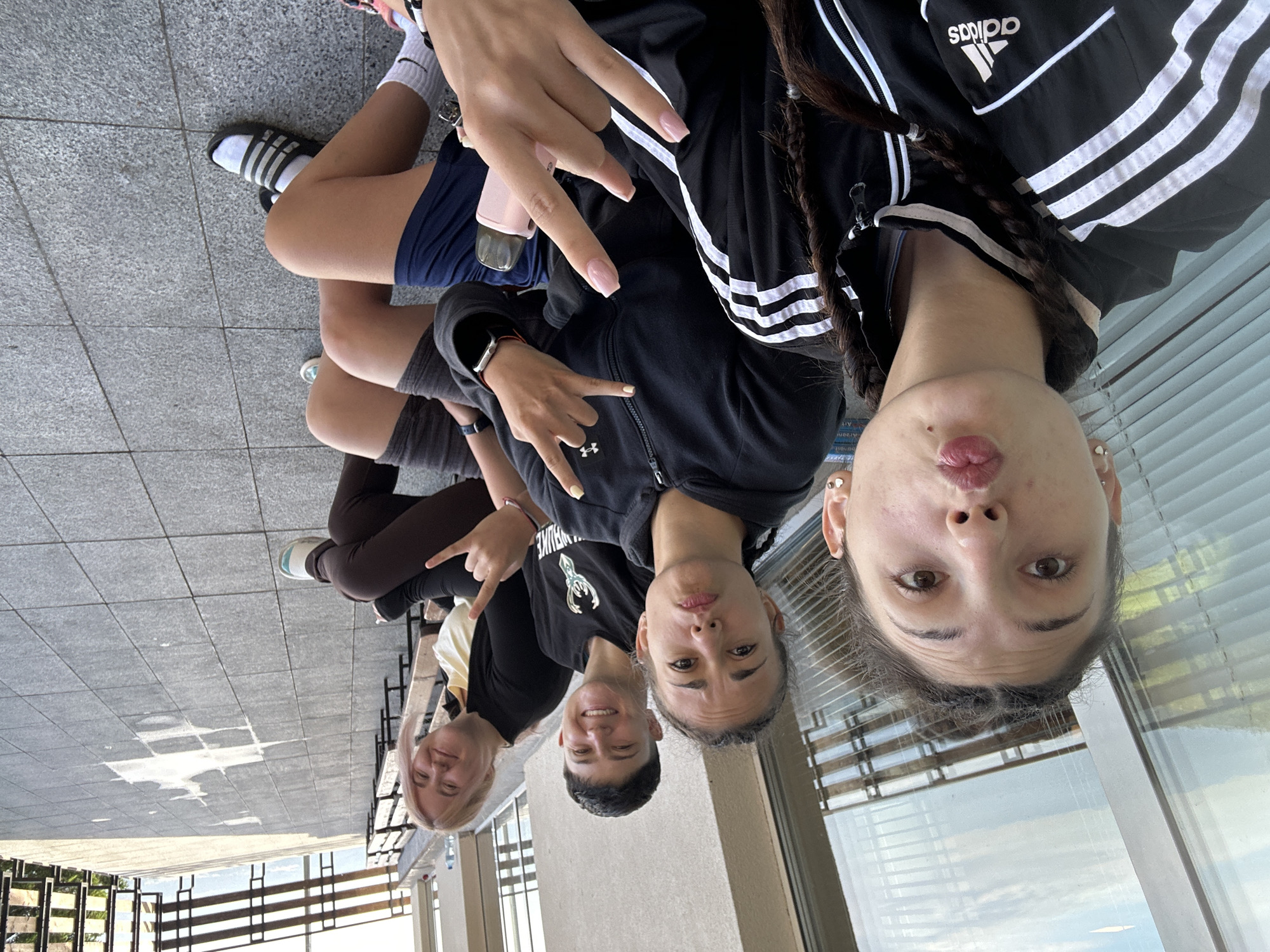 Four young women are seated on a bench outdoors on a paved area. The woman closest to the camera is in the foreground and is looking directly at the camera, puckering her lips. She is wearing a black and white Adidas track jacket and her hair is in two braids. To her right, a woman with short, dark hair is smiling and looking towards the camera. She is wearing a black t-shirt. Next to her, a woman with blonde hair is also smiling and looking at the camera, holding up her hand in a rock-on gesture. She is wearing a black jacket over a yellow shirt. The fourth woman is partially visible on the far right, also smiling. They are seated on a wooden bench with a railing behind them. In the background, there is a building with large windows and some vegetation. The ground is tiled with grey square tiles. The scene appears to be a casual gathering or hangout. It is not possible to determine the precise location within Bistritsa, Bulgaria, from the image alone, but the presence of outdoor seating, a building, and greenery suggests a public or recreational area.