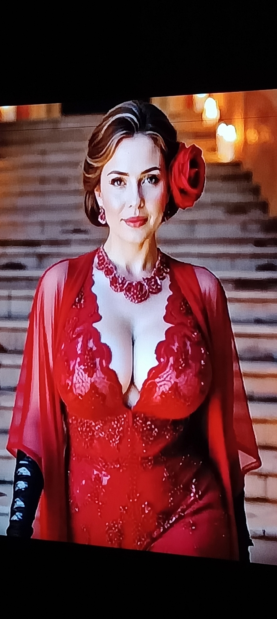 A woman with brown hair styled in an updo and a red rose accessory is seen from the chest up. She is wearing a low-cut red sequined dress with lace trim and a matching red beaded necklace. She also wears black gloves. The background appears to be a grand staircase with flickering lights. There is no identifiable information in the image to suggest it is located in Taganrog, Russia.