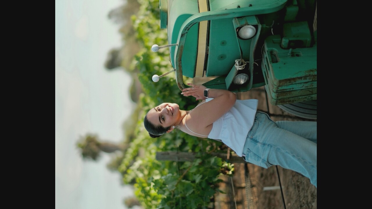 A young woman with dark hair pulled back in a bun poses next to a green tractor in a rural setting. She is wearing a white tank top and light-wash jeans, and she is leaning on the tractor with her hands. The tractor appears to be older, with some rust visible on its surface. The background consists of lush green foliage, likely a vineyard, and a light-colored sky. The overall lighting suggests it is daytime, possibly late afternoon due to the warm tones.