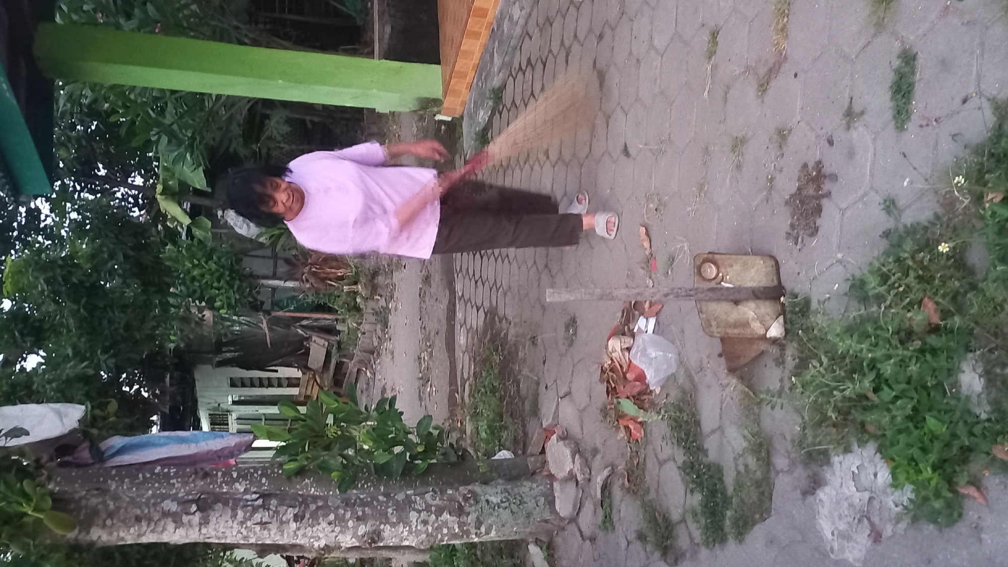 An outdoor scene at dusk in Pare, Indonesia depicts a woman sweeping a tiled walkway with a broom. The woman, with dark hair, wears a pink shirt and brown pants. She is positioned mid-stride, actively sweeping. The walkway is bordered by vegetation and a tree trunk on the left. To the right, a green pillar and a building are visible. Debris, including leaves and a plastic bag, is scattered on the ground. A rusty metal container is also present near the tree.