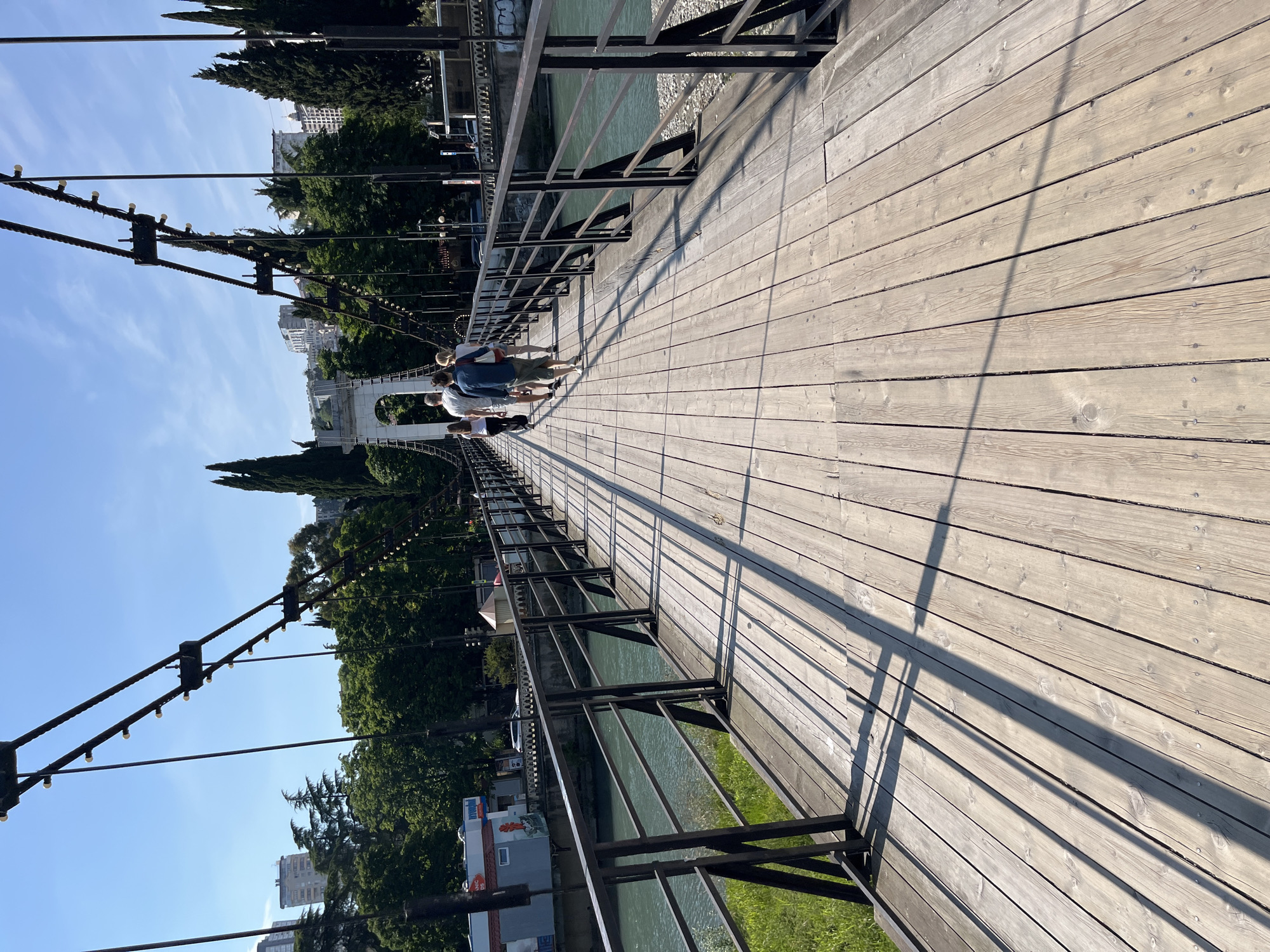 The image displays a pedestrian suspension bridge in Sochi, Russia, likely spanning the Sochi River near Riviera Park, under a clear blue sky. The sunlight is bright, creating long, distinct shadows of the bridge's structures across its wooden plank deck.
The bridge deck is made of light-colored, weathered wooden planks laid longitudinally, secured with visible fasteners. Black metal railings, comprising vertical posts and multiple horizontal bars, line both sides of the bridge, casting prominent diagonal shadows on the deck. Overhead, heavy dark suspension cables run parallel to the bridge, adorned with a series of small, illuminated light fixtures. Thinner vertical cables connect these main suspension lines to the bridge structure. In the distant center, a large, ornate white arched pylon supports the bridge, with similar structures implied beyond the frame.
A group of approximately five to six individuals, appearing to be adults, is walking away from the viewer along the bridge towards the distant arch. From left to right in the primary group, a woman in dark attire, a man in a light t-shirt and dark shorts, a man in a blue long-sleeved shirt and light shorts, and a woman in a white top carrying a bag are visible. They are dressed in casual summer clothing.
To the left of the bridge, a body of turquoise-colored water, consistent with a river, flows below. The far bank of the river shows sparse vegetation and some rocky terrain. The background reveals a landscape of dense green trees, including tall, slender cypress-like conifers, interspersed with numerous multi-story buildings on rising terrain. On the immediate left bank of the river, beneath the bridge, a small commercial building with a "Hyundai" sign is visible, along with other urban infrastructure and vehicles.