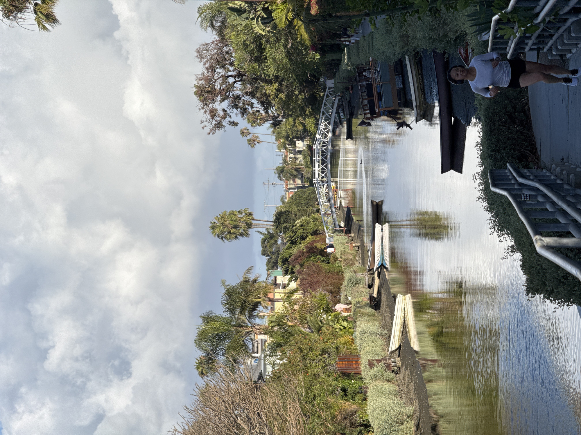 The image captures a view of the Venice Canals in Los Angeles, California. A woman in athletic wear is jogging along a concrete path that runs alongside the canal. The canal itself is narrow and filled with water, reflecting the sky and surrounding foliage. Various small boats and wooden structures line the edge of the canal. Lush greenery, including trees and shrubs, is visible on both sides of the waterway, with residential houses partially visible amidst the vegetation. A pedestrian bridge spans the canal in the distance. The sky above is overcast with clouds.