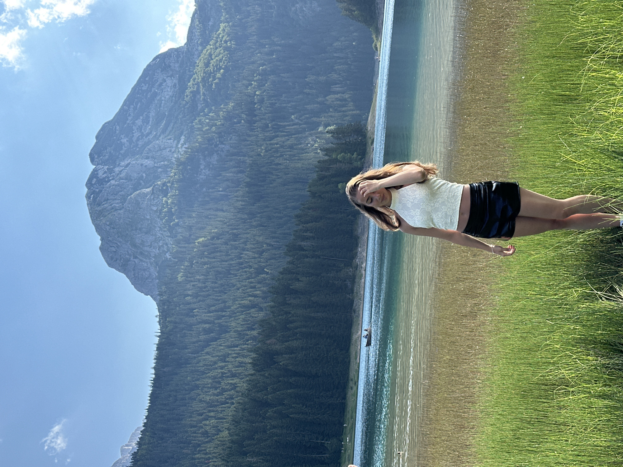 A young woman with long, wavy brown hair stands in a field of tall green grass, facing slightly to her right. She is wearing a white lace halter top and black shorts. Her right hand is raised to her forehead, and her eyes are closed. Behind her, a clear blue lake stretches out, with a small boat visible on its surface. Beyond the lake, a dense forest of dark green pine trees covers a steep mountainside, which rises to a rugged, rocky peak under a bright blue sky with a few wispy clouds. The scene is set in Pitomine, Montenegro.