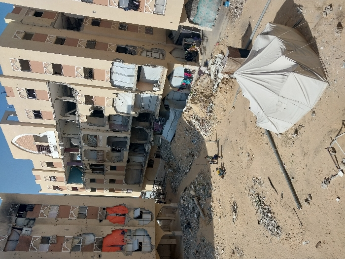 An aerial perspective of a severely damaged urban landscape in Khan Yunis, Palestinian Territory, depicts a scene of extensive destruction. Three multi-story residential buildings, constructed with light-colored masonry and featuring patterned facade elements, show widespread structural and superficial damage. The central building exhibits the most significant devastation, with multiple floors partially collapsed or entirely exposed to the elements, revealing destroyed interiors. The building on the left has large sections of its facade missing, with bright orange tarpaulins covering some openings and numerous bullet or shrapnel impacts visible across its surface. The building to the right also displays broken windows and exposed rooms.
The ground in the foreground and midground consists of barren, sandy earth mixed with scattered concrete debris, rebar, and large piles of rubble. In the midground, near one such pile, four individuals are gathered, indistinct in their specific actions due to distance, with a yellow object beside them. A long, dark pipe or conduit traverses the lower midground. In the lower right foreground, a large, light-colored canvas tent, possibly a temporary shelter, is erected on the sandy ground, secured with ropes and stakes. The sky above is clear and bright.