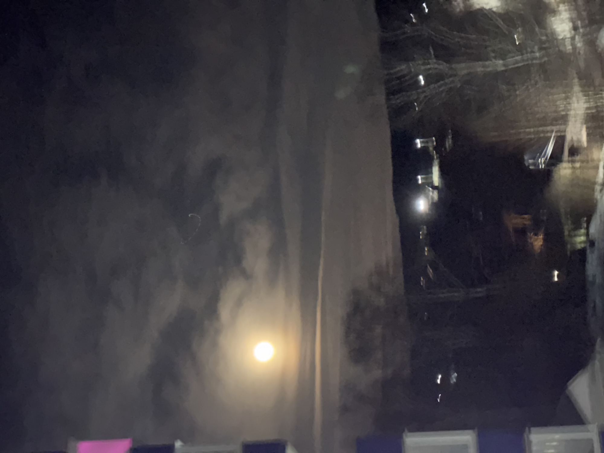 A nighttime shot depicts a full moon shining brightly through the clouds, partially obscuring its light. The moon is positioned above a building, but the location or city is not evident from the image alone, and cannot be determined using only forensic means. The building is shrouded in darkness, with only a few lights visible. The foreground contains indistinct forms, which may be rooftop structures.