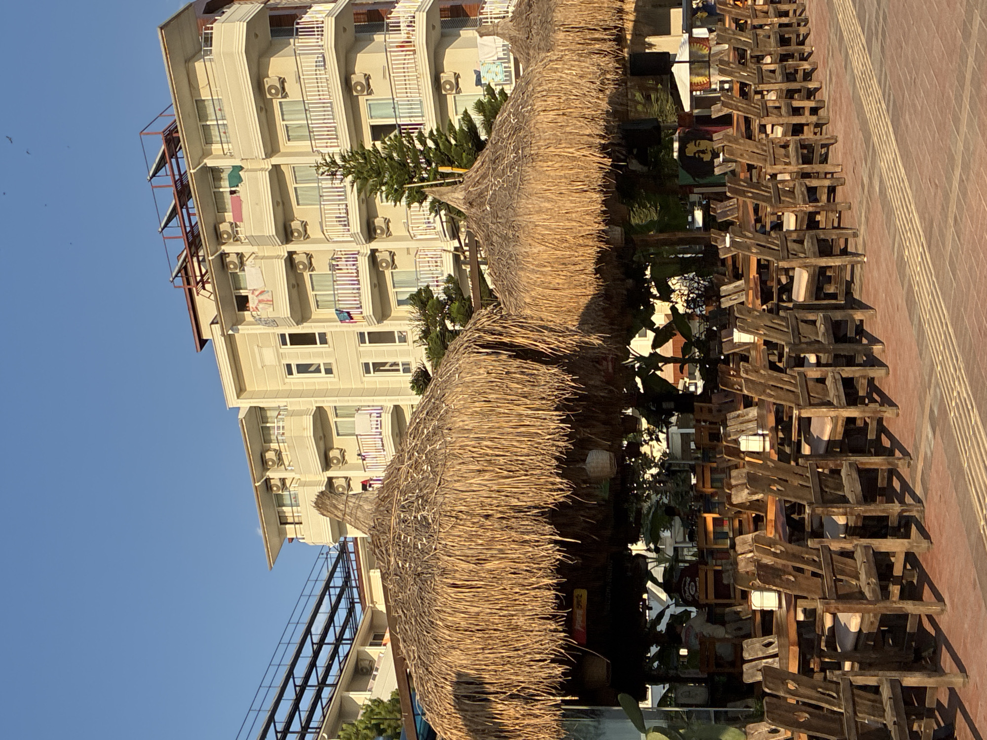 This is a photograph of an outdoor seating area for a restaurant or cafe in Alanya, Turkey. The scene is dominated by several large, thatched umbrellas providing shade over rows of wooden tables and chairs. In the background, a modern, light-colored apartment building rises several stories high, with balconies and air conditioning units visible. The sky is clear and blue. The ground in front of the seating area is paved with red brick. The overall impression is of a casual, sun-drenched environment, possibly a tourist area.