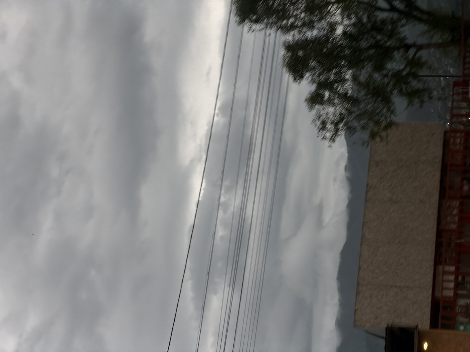 The image shows a cloudy sky with power lines running across the frame. A tree is visible on the right side of the image, next to a building under construction or renovation. The location is suspected to be Tbilisi, Georgia.