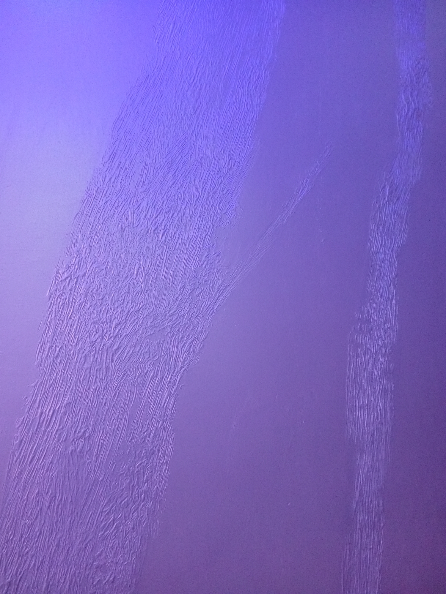 The image displays a close-up, abstract view of a textured purple wall. The wall has a rough, uneven surface with visible striations and indentations that create a pattern of light and shadow. The lighting appears to be artificial, casting a purplish hue across the scene. There are no people, animals, or discernible objects in the frame. The background is a solid, textured purple surface. This image does not provide any information or visual cues that would allow for its location to be identified in Sabon Gari East, Nigeria, or any other specific geographical area.
