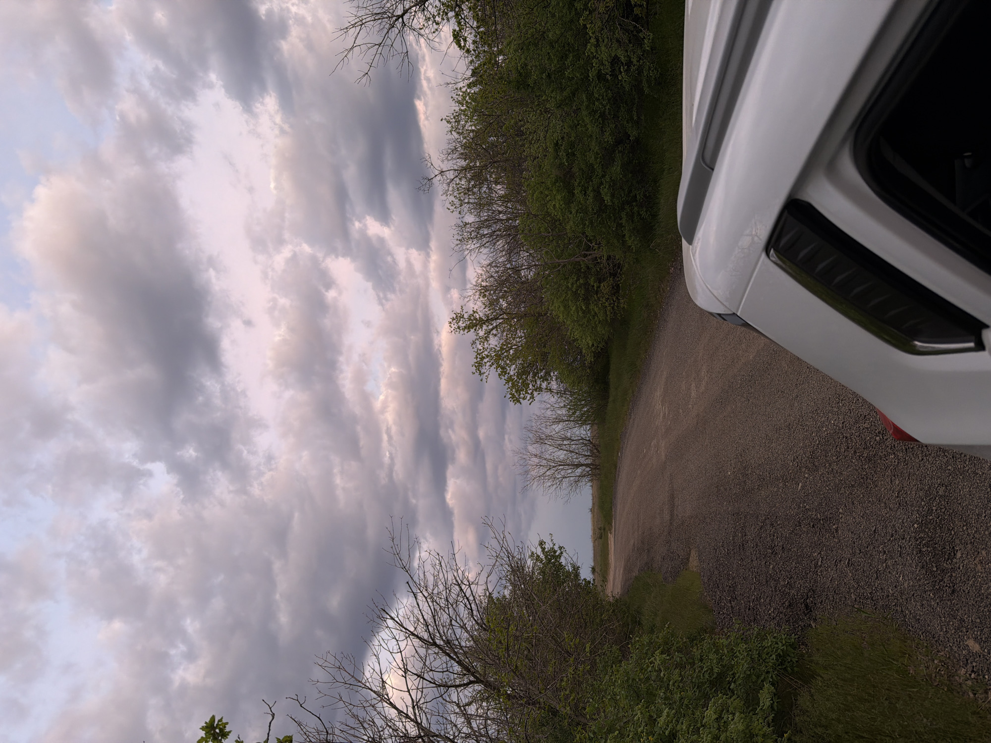 The image depicts a gravel road curving away into the distance. Lush green vegetation lines the sides of the road. A white vehicle is parked on the right side of the frame, obscuring part of the scene. Above, the sky is filled with clouds, ranging in color from light gray to dark gray. The image appears to be taken at dusk or dawn, as the light is soft. The location is identified as Ternovka, Russia.
