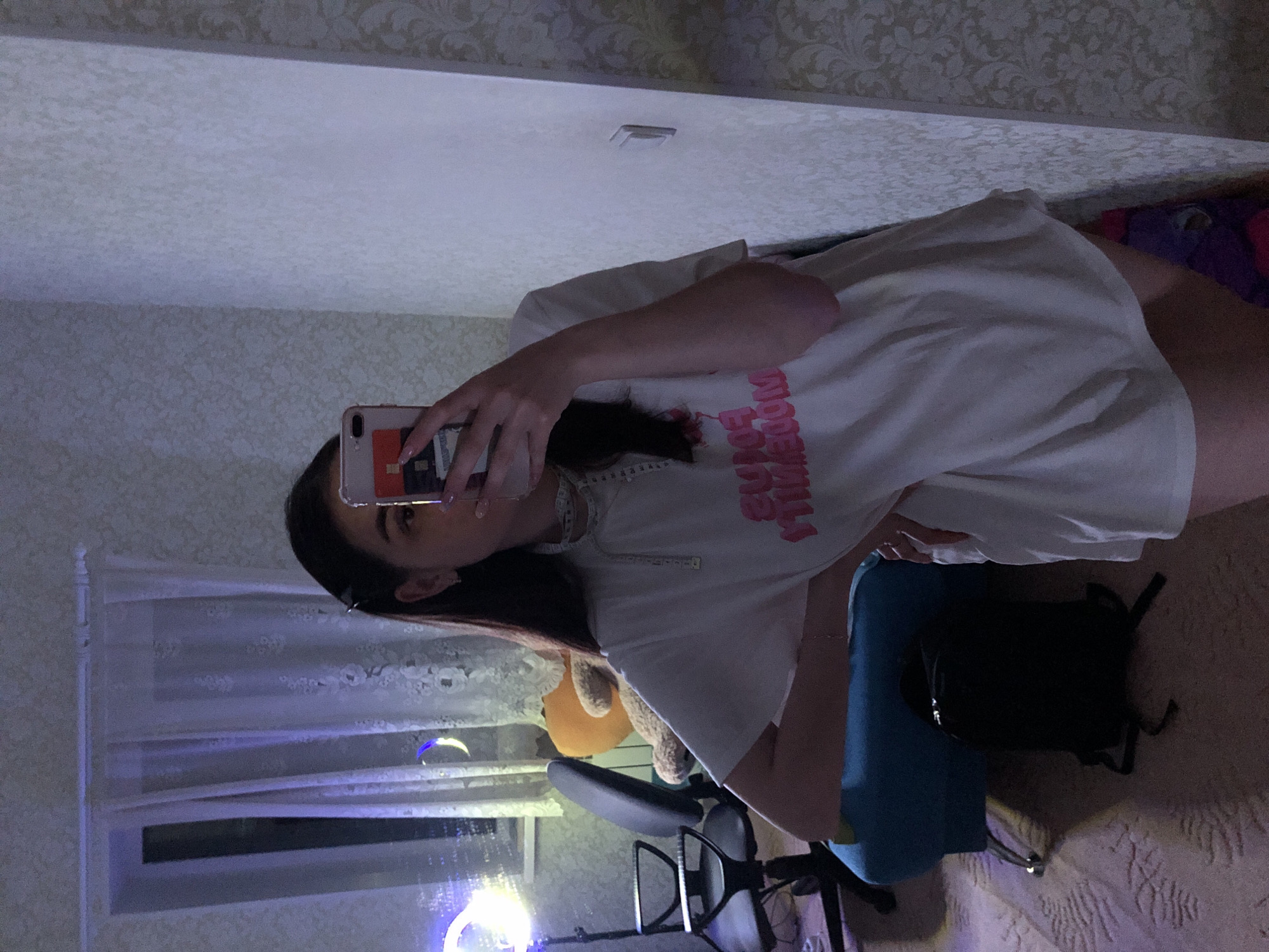 A woman is taking a selfie in a room. She is wearing a white t-shirt with pink lettering. Her hair is dark, and she is holding a cell phone. Behind her is a window with sheer curtains. There is a desk and chair in the room. The wallpaper has a floral pattern. A teddy bear is visible in the background. The location may be in Noviy, Russia.