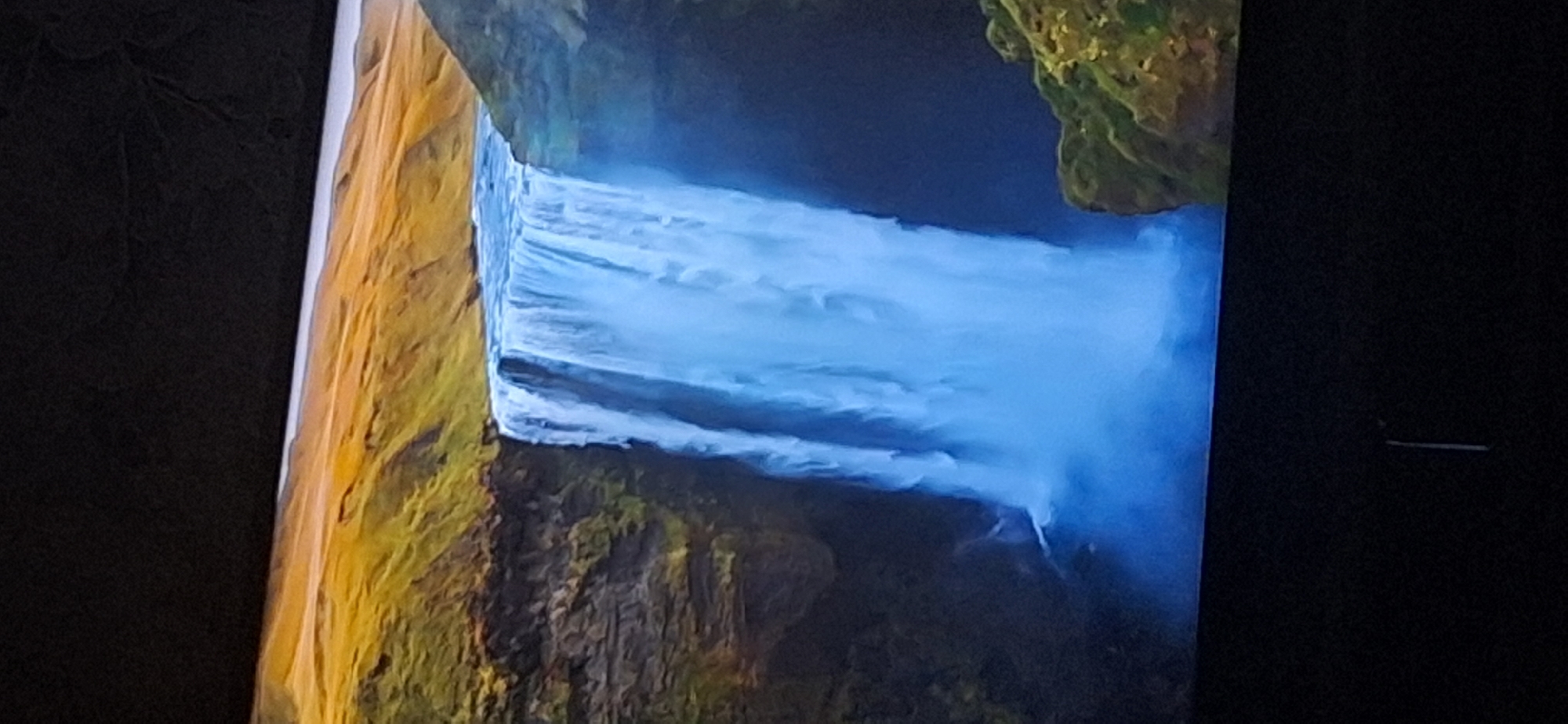 The image shows a majestic waterfall cascading over a cliff face. The water is a vibrant blue, creating a misty spray at the base. The surrounding landscape is characterized by rolling hills covered in green and yellow vegetation, with darker rock formations visible on the cliff. The sky above is a dark, undefined expanse. There are no people or man-made structures visible in the scene. The location of this waterfall is not in Bila Tserkva, Ukraine.