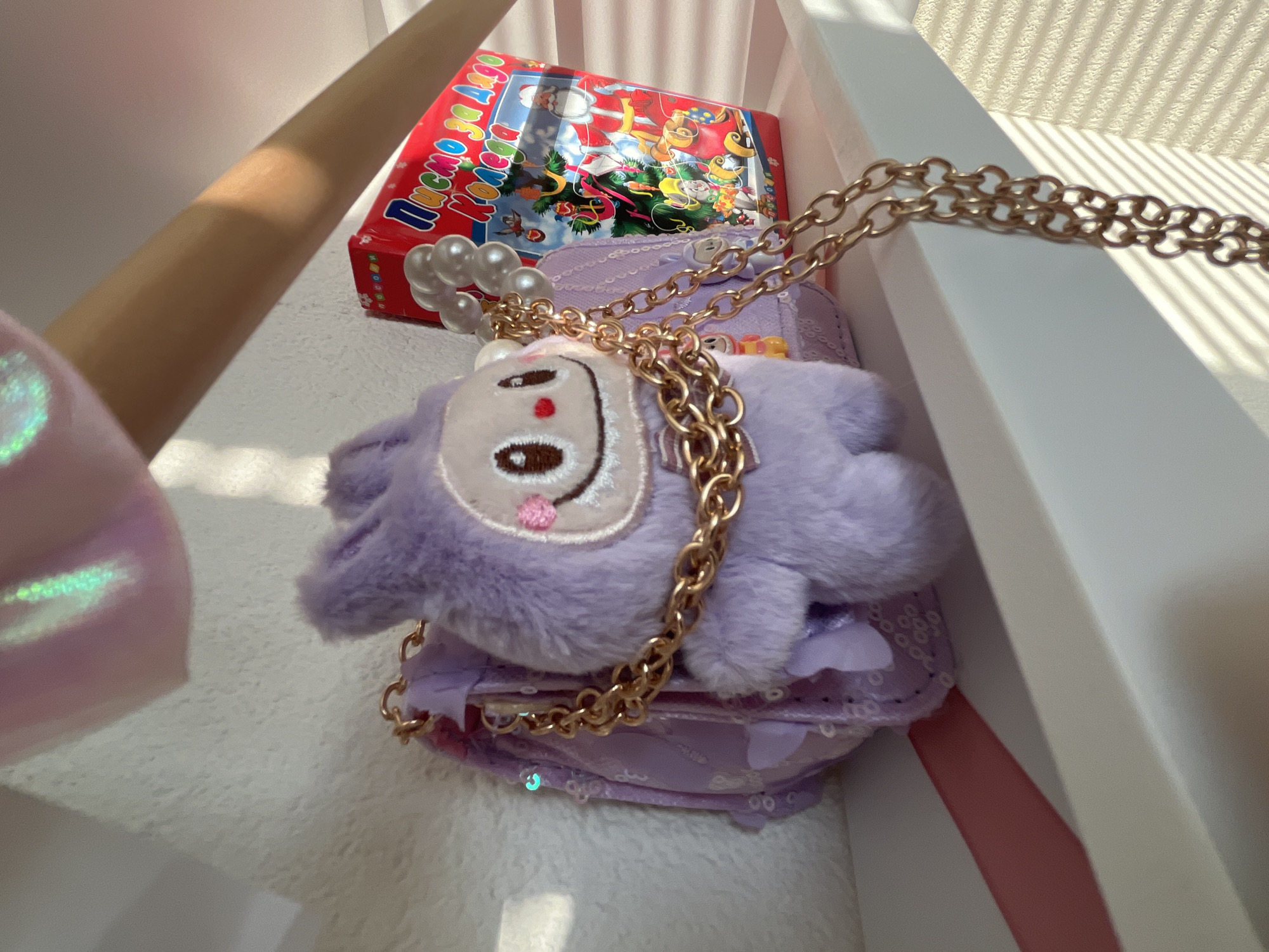 A close-up photograph shows a purple, plush purse with a cartoon face detail, adorned with a gold chain and faux pearl accents. The purse is positioned on a white surface with a red box placed behind it. The scene is lit by natural light, casting shadows from the sun.