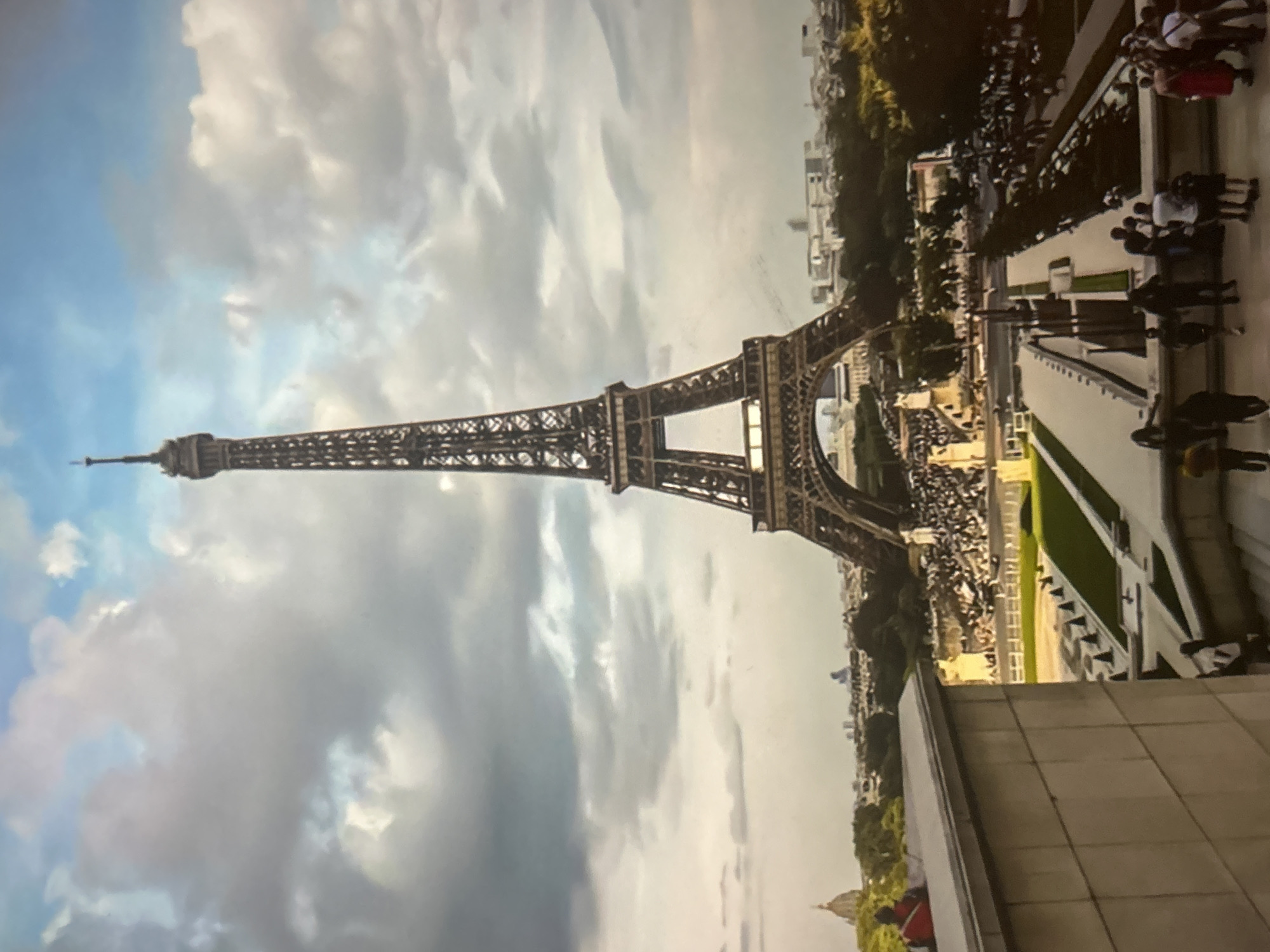 The image shows the Eiffel Tower in Paris, France, under a cloudy sky. In the foreground, there is a modern building with a tiled facade. Below this, there are manicured gardens with water features and rows of dark cones. A crowd of people is visible in the middle ground, gathered around the base of the Eiffel Tower and in the gardens. The Eiffel Tower itself is the dominant feature, with its intricate iron structure reaching towards the sky. The background shows a cityscape with trees and other buildings, including a domed structure on the left. Several people are scattered throughout the scene, some walking, some standing, and some appearing to be observing the surroundings. The overall impression is a bustling tourist scene in front of a world-famous landmark.