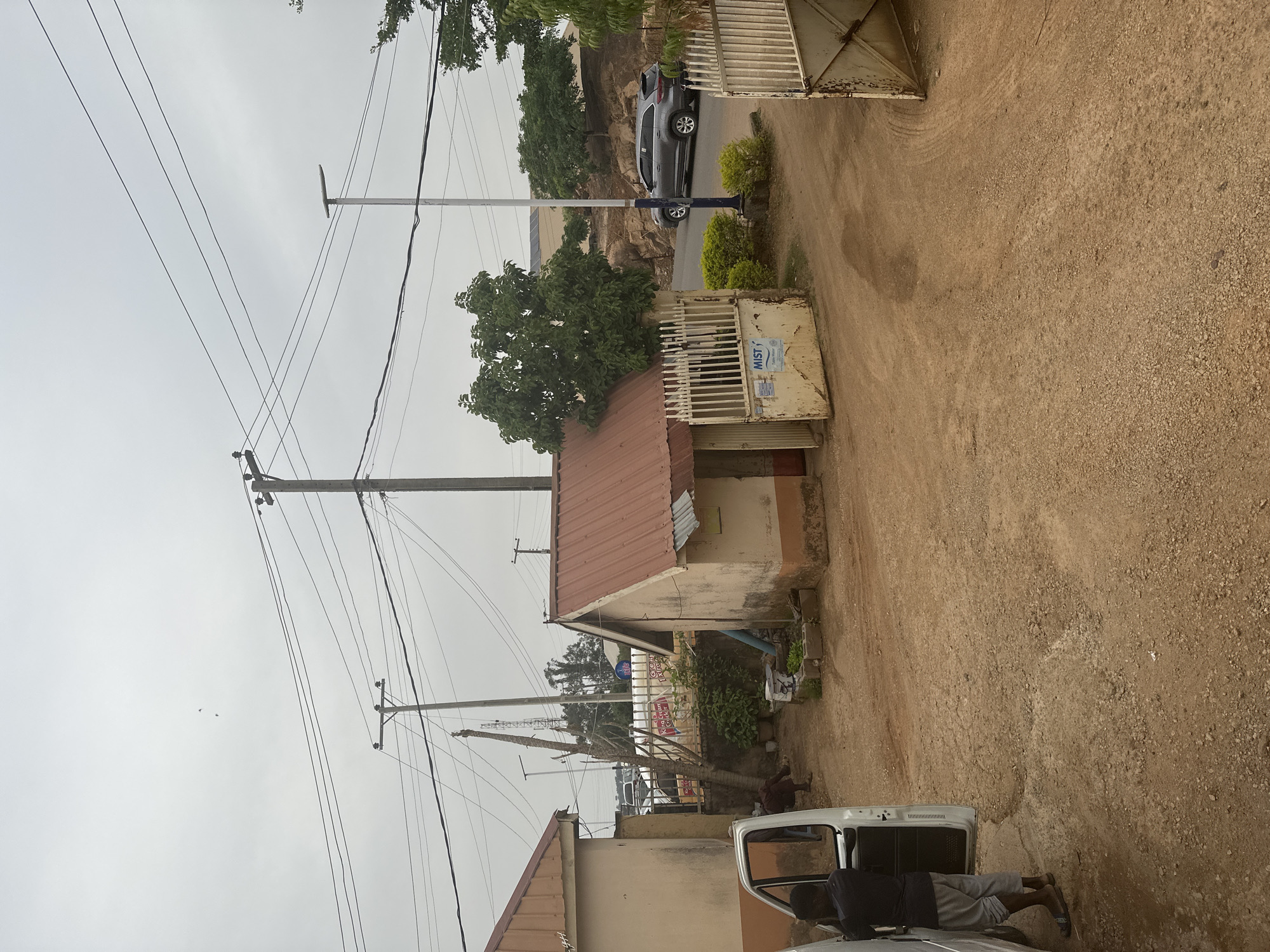 This image captures a street scene in what appears to be a semi-urban or rural area of Jos, Nigeria, under an overcast sky. The ground in the immediate foreground is unpaved, consisting of light brown dirt and gravel.
In the lower left, a white vehicle, possibly a van or pickup truck, is parked with its passenger door open. A person is partially visible inside or near the open door, wearing shorts and a dark top. Further into the scene, a series of small, single-story buildings with reddish-brown corrugated metal roofs and light-colored, somewhat weathered walls line what appears to be a compound or street.
One of these buildings features a white metal gate that is slightly ajar, revealing a sign behind it partially obscured by a tree, with visible text including "...King... Cream Chicken..." and "...Gen Express...". Another white metal gate, closer to the center, has a sticker or sign that reads "MUST..." along with "Sales Points" and other smaller, illegible text. Near this gate, a person is seen sitting on the ground.
A paved road runs through the middle ground, where a dark grey SUV is visible, either parked or moving slowly. Another person is standing next to this SUV. Beyond the road, the terrain rises, revealing rocky formations and dense green foliage from various trees.
The upper portion of the image is dominated by a light grey, overcast sky, indicating a cloudy day. Numerous utility poles stand tall, supporting a complex web of overhead power lines that crisscross the frame. The overall atmosphere suggests a normal daytime setting with diffuse natural light.