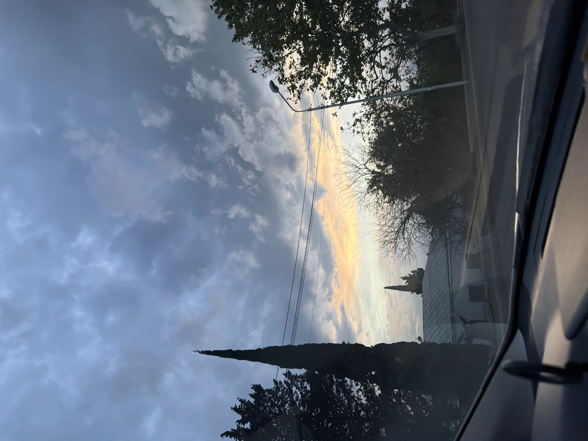 The image is a low-angle shot looking up through a car windshield. The view includes a sky with a mix of dark, overcast clouds and patches of light, possibly from a setting sun. There's a single lamppost visible, with bare branches of trees silhouetted against the lighter parts of the sky. Power lines run across the frame. A tall, thin cypress tree and another tree with darker foliage are in the foreground. In the middle ground, a white building with a pointed spire on the roof is visible. The scene is likely in Tbilisi, Georgia, given the provided context. The image captures a transitional weather moment during either sunrise or sunset, viewed from inside a vehicle.