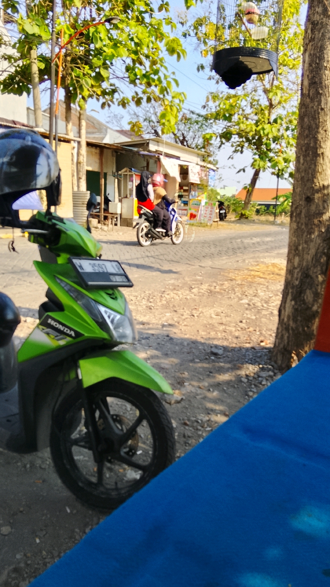 In Dahanrejo, Indonesia, a bright green Honda scooter is parked on a dusty road, its front wheel angled to the left. The scooter is in the foreground, and its license plate reads "W 6597 LN". Behind the scooter, a person rides another motorcycle, wearing a red jacket and dark pants. They are captured mid-motion, moving away from the camera.
The scene is set on a road that appears to be unpaved, with gravel and dirt underfoot. To the left of the road, there are small shops or stalls with corrugated metal roofs. Trees with lush green leaves line the background, their branches casting shadows. One tree on the right side of the image has a thick trunk and is in sharp focus. Above the shops, a birdcage hangs from a tree branch, containing at least two birds. The overall atmosphere suggests a warm, sunny day in a semi-urban or rural Indonesian setting. The foreground is partially obscured by a vibrant blue surface, possibly a mat or fabric, angled across the bottom of the frame.