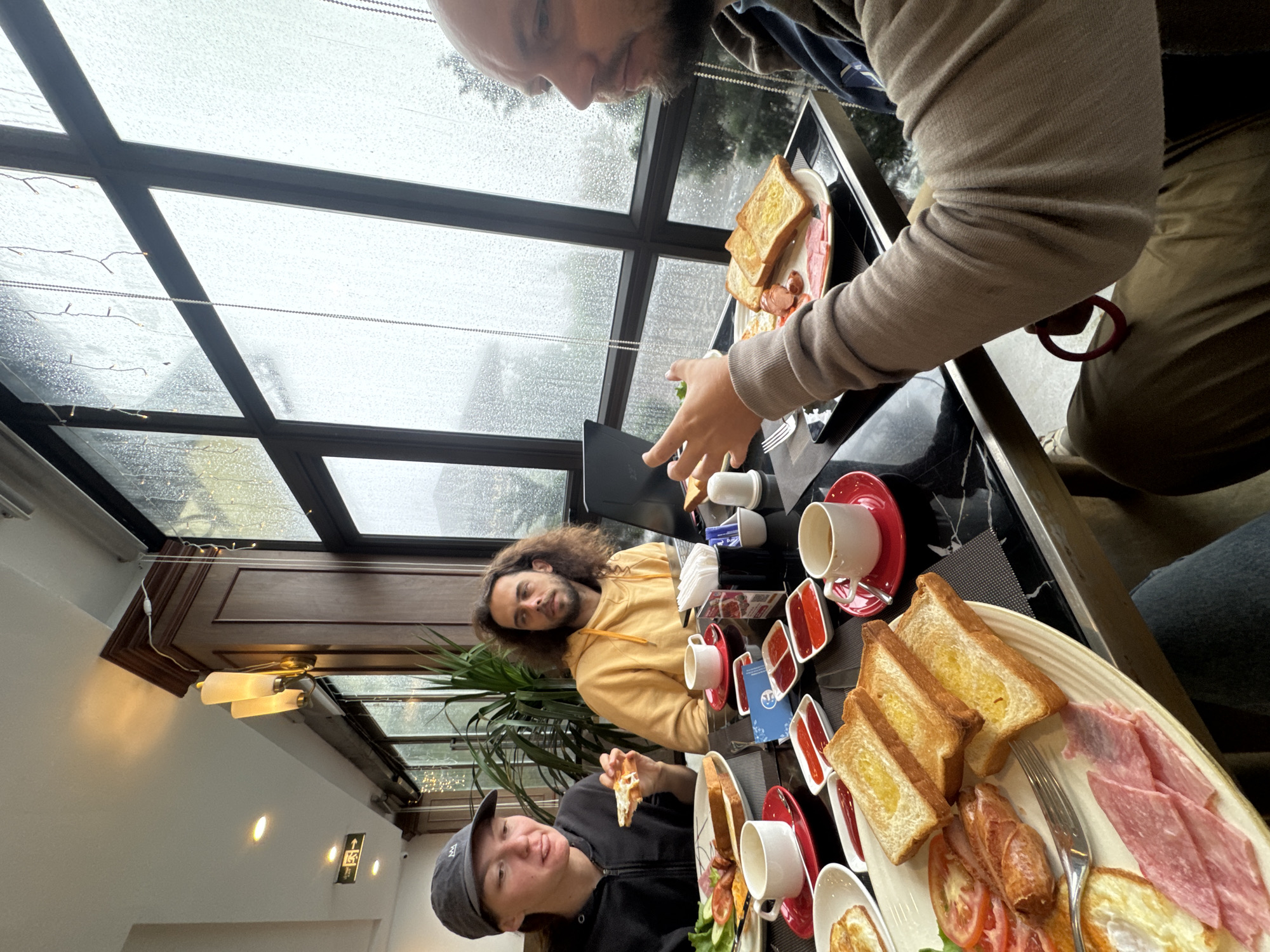 Three people are seated around a black marble table laden with breakfast food, including toast, ham, sausage, eggs, tomatoes, lettuce, and small jars of what appears to be jam. Coffee cups and saucers are also present. The people are casually dressed. A man with a beard and tan sweater is standing and pointing at something on the table. A man with long brown hair and a yellow hoodie is seated, looking at the camera. A woman with a baseball cap and black jacket is eating a piece of toast. The background shows a window with a view of a cloudy or rainy day. The location is indoors, with interior lighting fixtures visible. The presence of rain and the style of architecture point to the possibility of this photo being taken in Binh Lu, Vietnam.