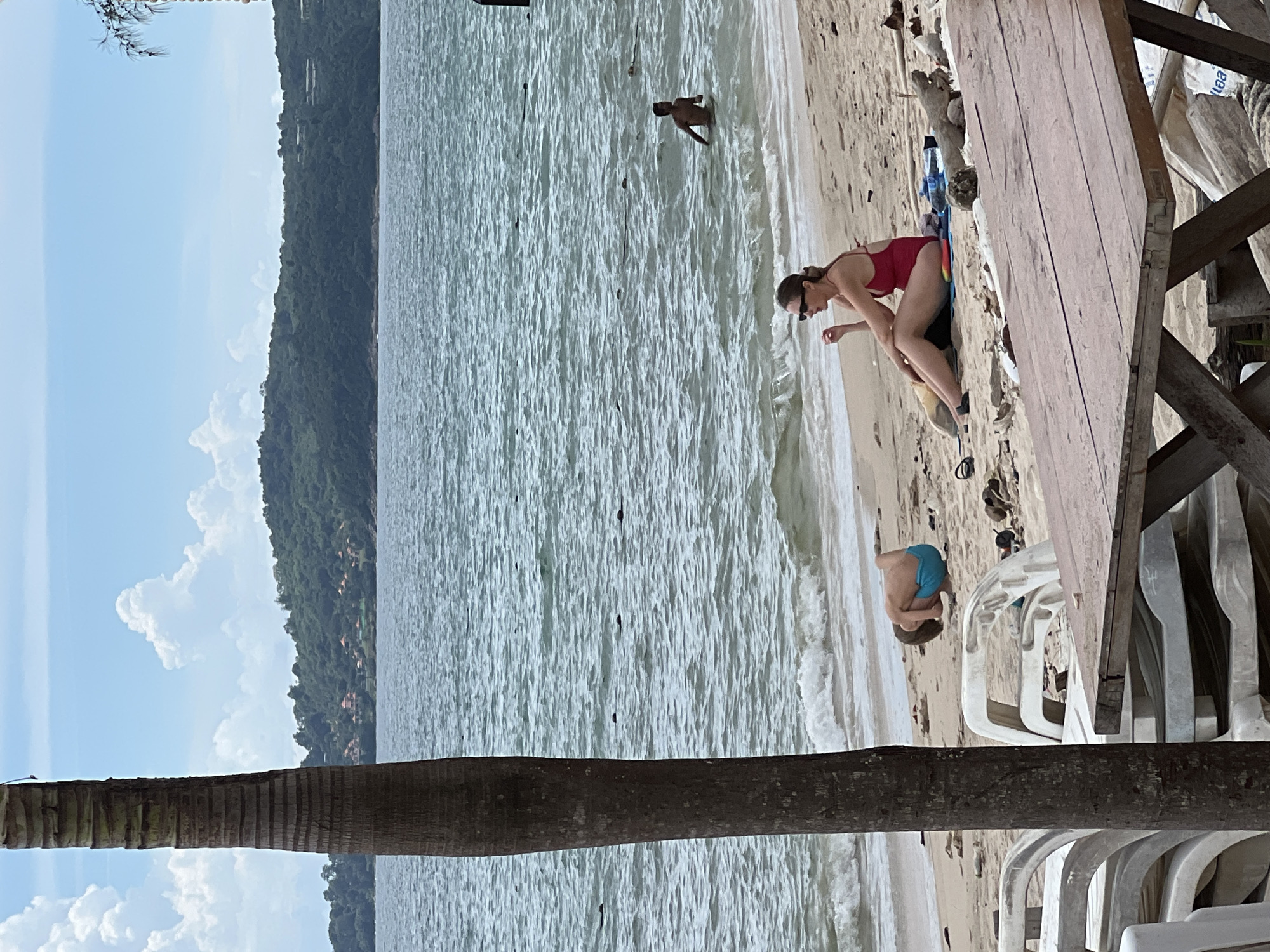 A medium shot captures a beach scene with a prominent palm tree trunk on the left. In the foreground, a rustic wooden picnic table is partially visible. On the sand, a woman in a red swimsuit sits facing left, wearing sunglasses, and appears to be talking. Beside her is a blue mat and some water bottles. Further back, a child in blue swim trunks is crouched down on the sand. In the ocean, a person is wading in the water, and several other dark, indistinct objects are visible in the water. The background features a lush, green hillside with scattered buildings and a bright blue sky with scattered clouds. The water is choppy, and small waves are breaking on the shore. White plastic lounge chairs are stacked near the foreground.